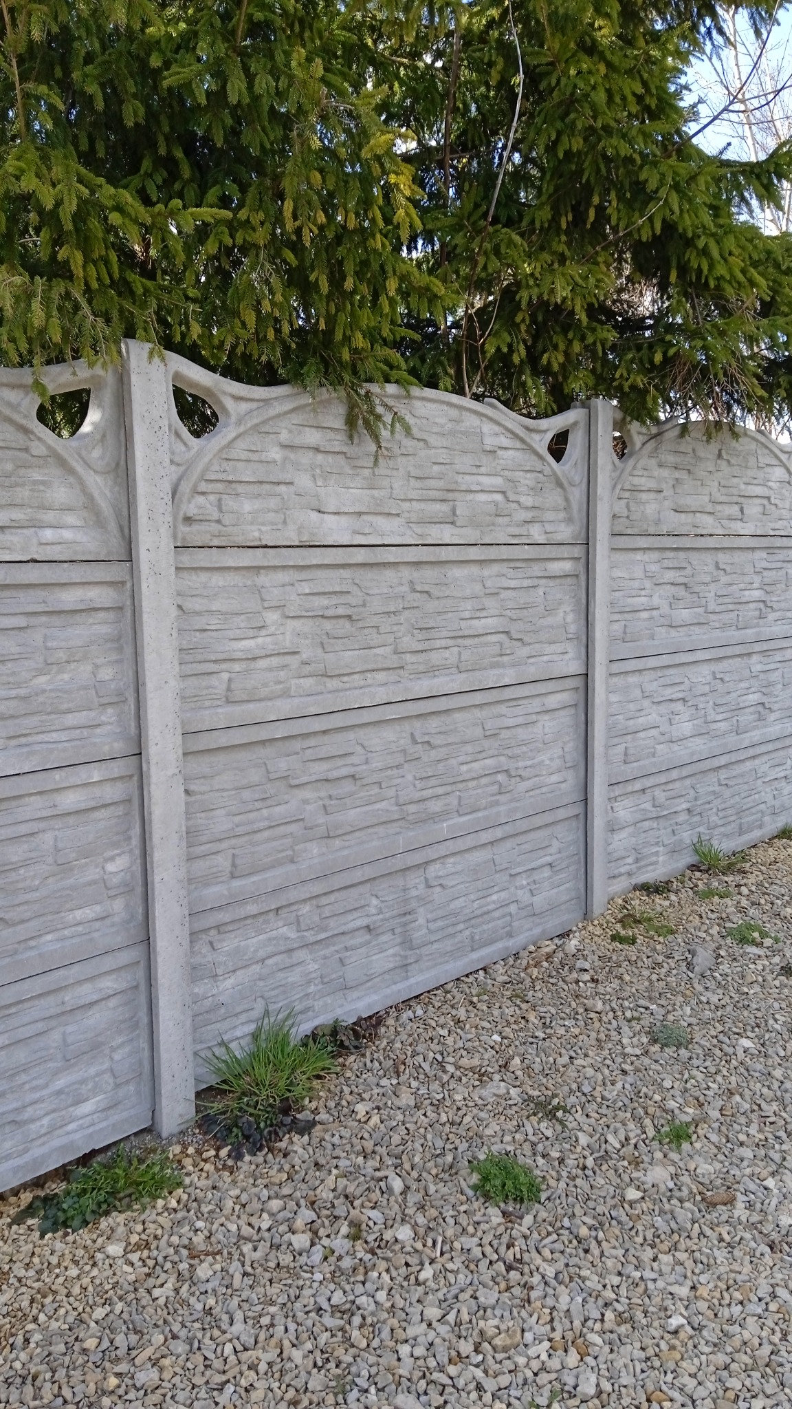 Szary, prefabrykowany płot z betonu imitującego kamień, z dekoracyjnymi wycięciami w górnej części, otoczony żwirem i zielenią, z iglastym drzewem w tle.