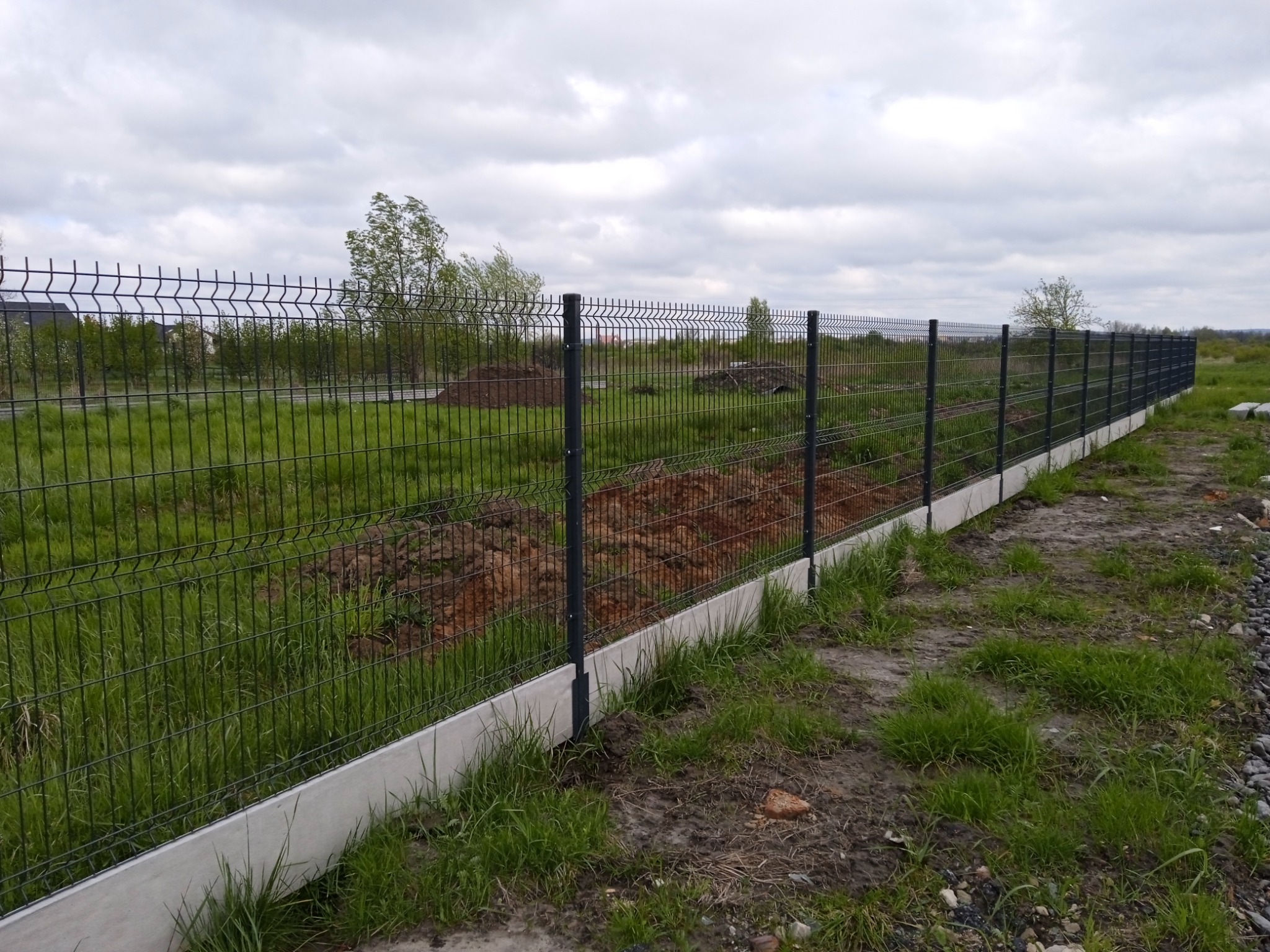 Długie, proste ogrodzenie panelowe z podmurówką betonową, widoczne na tle łąki i pochmurnego nieba.