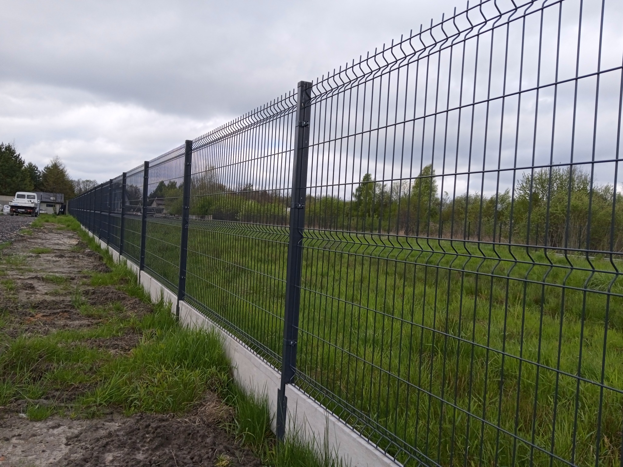 Długie, proste ogrodzenie panelowe w kolorze grafitowym, osadzone na betonowej podmurówce, rozciągające się wzdłuż zielonej trawy i grząskiej ziemi, z widocznym samochodem dostawczym w oddali.