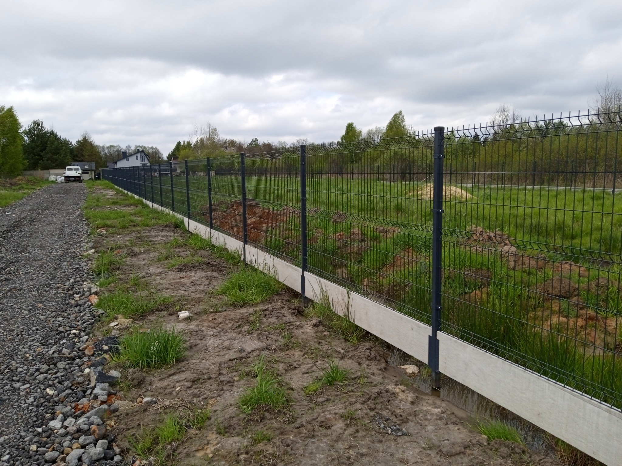 Długi, ciemny panelowy płot z podmurówką z jasnego betonu na nierównym terenie z widokiem na pole i drzewa w tle.