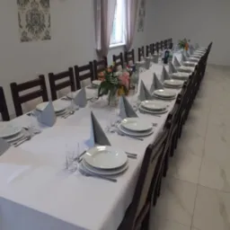 Długi stół nakryty białym obrusem, zastawiony talerzami, sztućcami, szklankami i serwetkami, udekorowany kwiatami, przygotowany na uroczystość, z balonami w kolorach czarnym, białym i złotym...