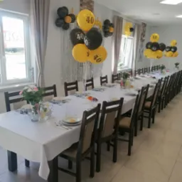 Uroczysta sala z długim stołem nakrytym białym obrusem, dekoracje z czarno-żółtych balonów z cyfrą 40, kwiaty w wazonach, zastawa stołowa dla gości. Przygotowania do przyjęcia urodzinowego.