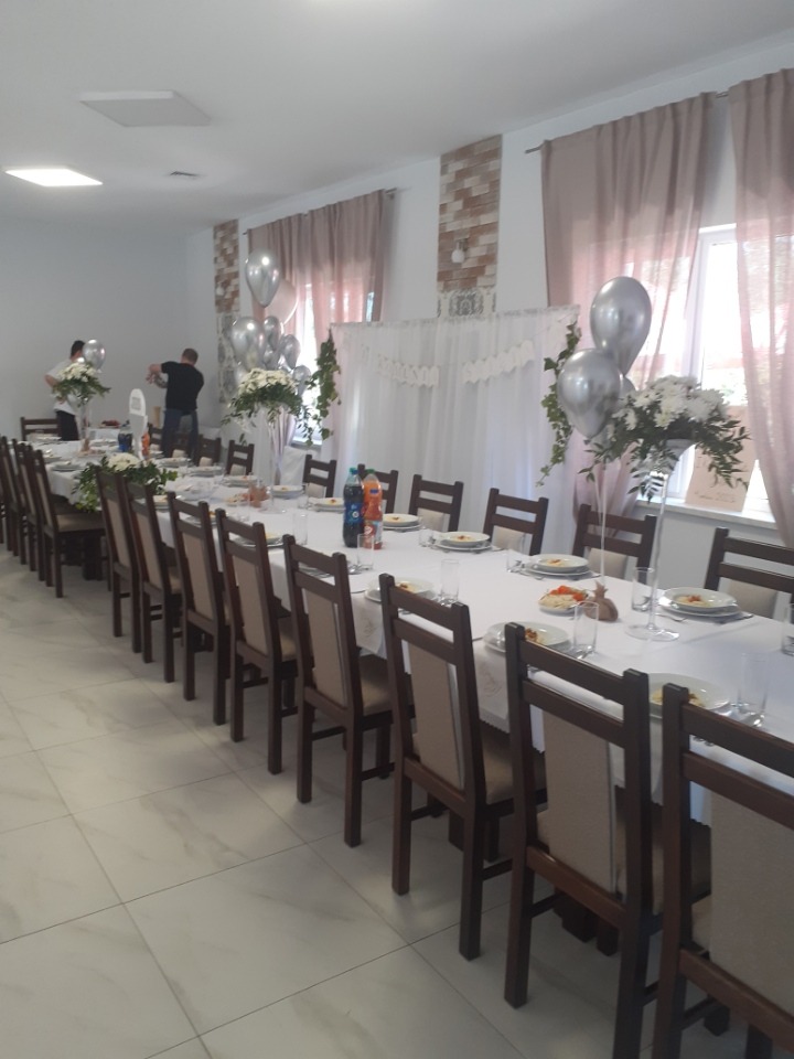 Długi stół nakryty białym obrusem, ustawiony w sali z jasnymi ścianami i podłogą, przygotowany na uroczystość. Dekoracje z białych kwiatów i srebrnych balonów. W tle osoby przygotowujące salę.