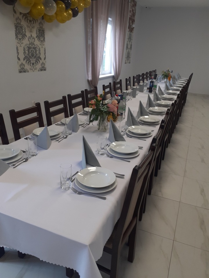 Długi stół nakryty białym obrusem, zastawiony talerzami, sztućcami, szklankami i serwetkami, udekorowany kwiatami, przygotowany na uroczystość, z balonami w kolorach czarnym, białym i złotym...