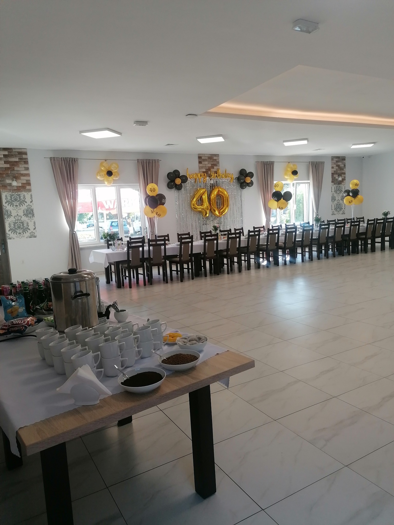 Sala bankietowa przygotowana na przyjęcie urodzinowe, stoły nakryte białymi obrusami, dekoracje z balonów w kolorach czarnym i złotym, na stole z przodu naczynia i napoje.