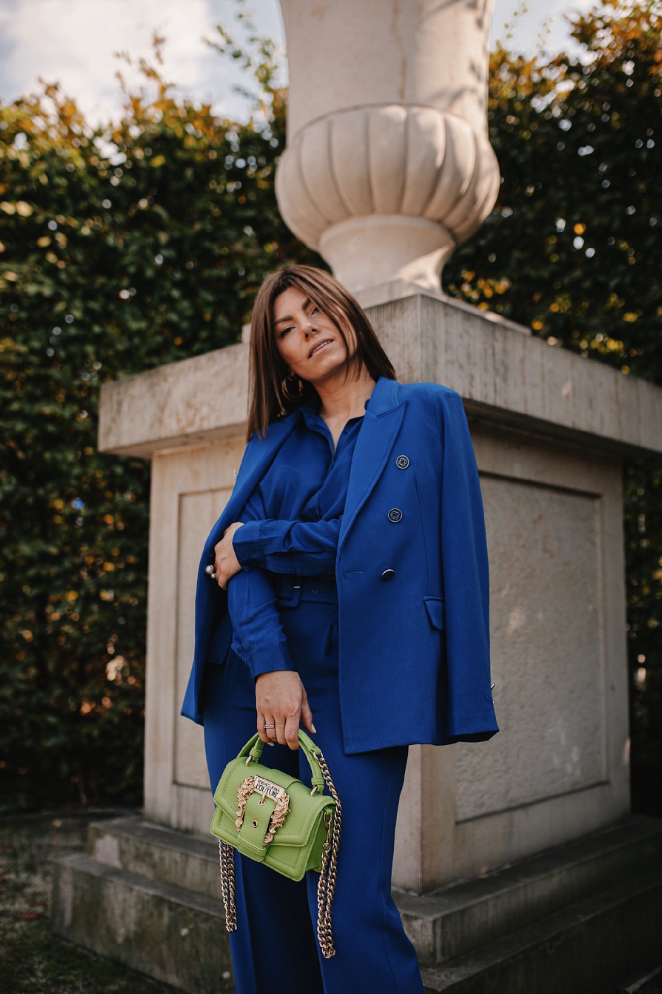 Kobieta w kobaltowym garniturze pozuje z limonkową torebką na tle kamiennej wazy i zieleni.