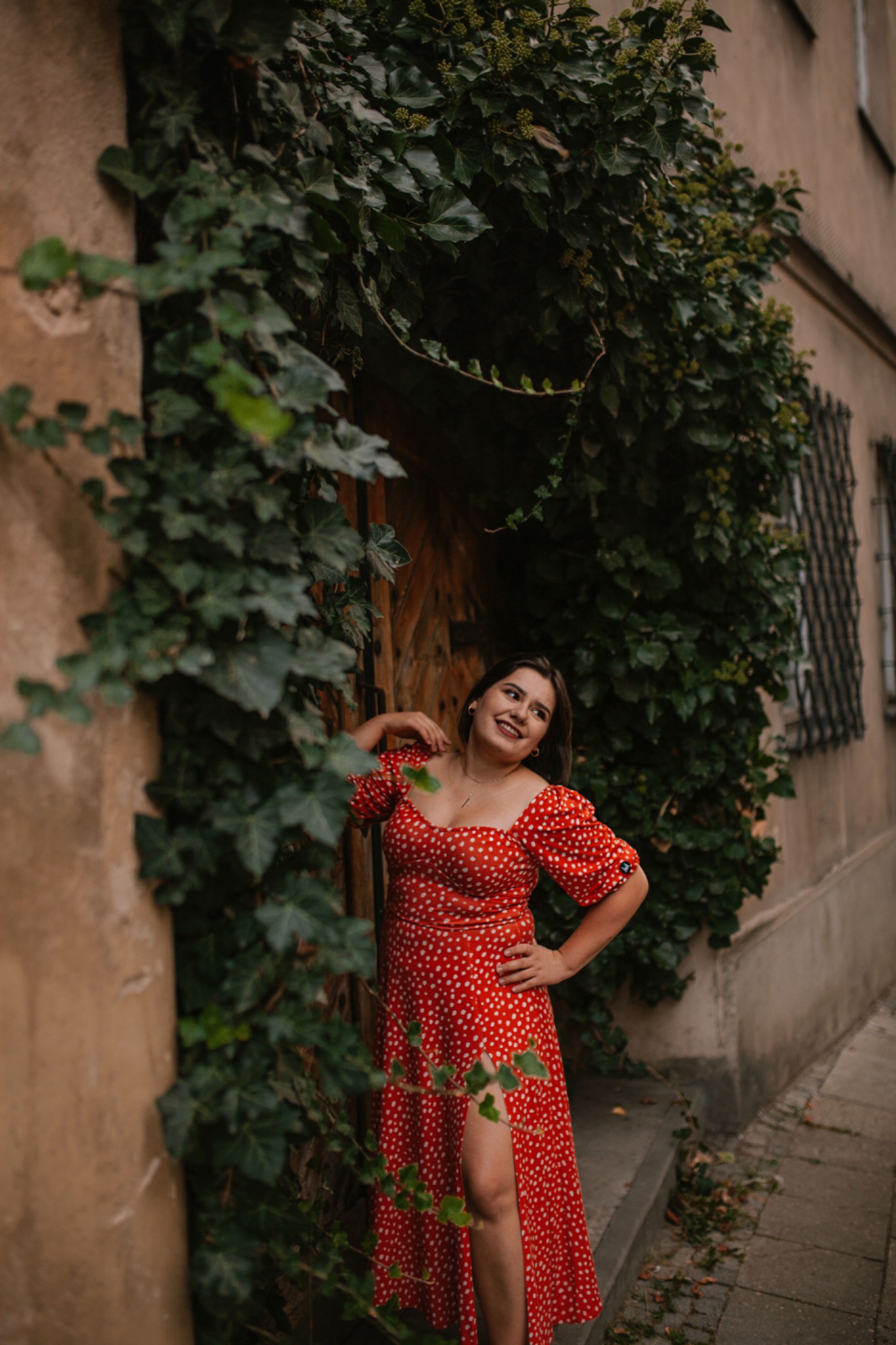 Uśmiechnięta kobieta w czerwonej sukience w białe kropki pozuje na tle ściany porośniętej bluszczem i drewnianych drzwi.