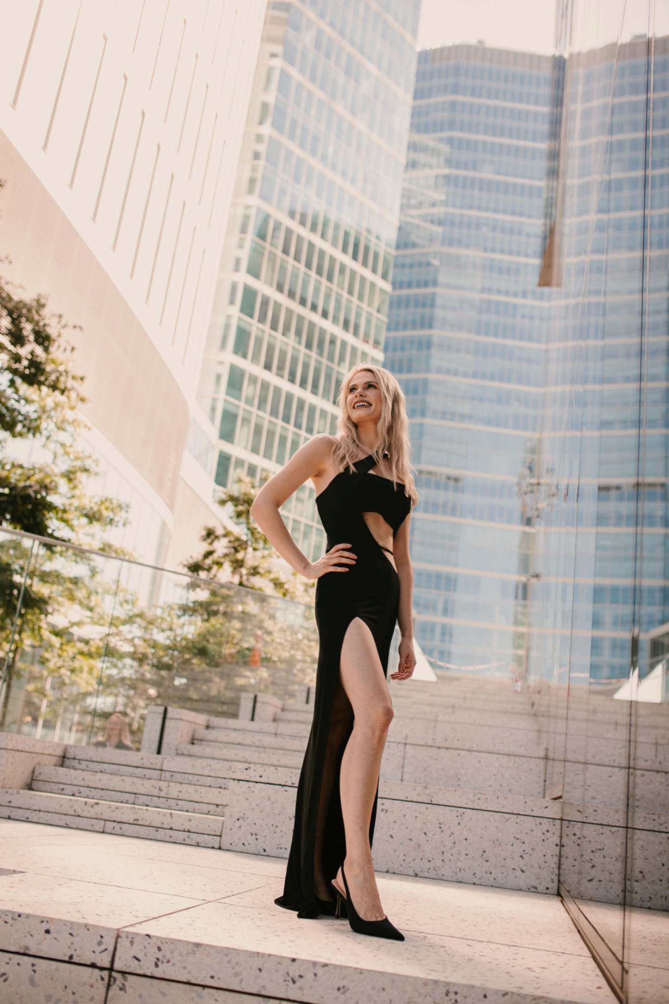 Kobieta w czarnej sukni z rozcięciem, pozująca na tle nowoczesnej architektury miejskiej, z uśmiechem spoglądająca w górę.