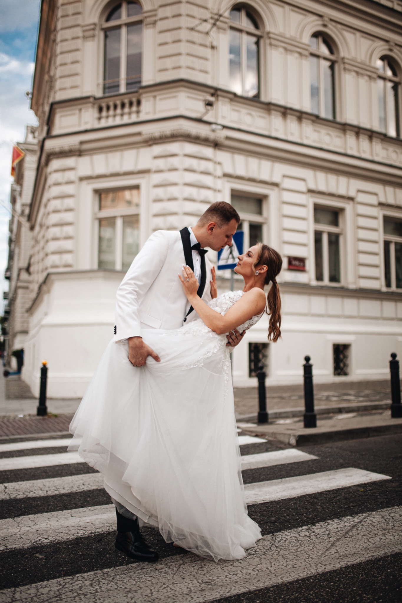 Para młoda w białych strojach, on w smokingu, ona w sukni ślubnej, obejmująca się na pasach dla pieszych przed budynkiem z jasną elewacją.