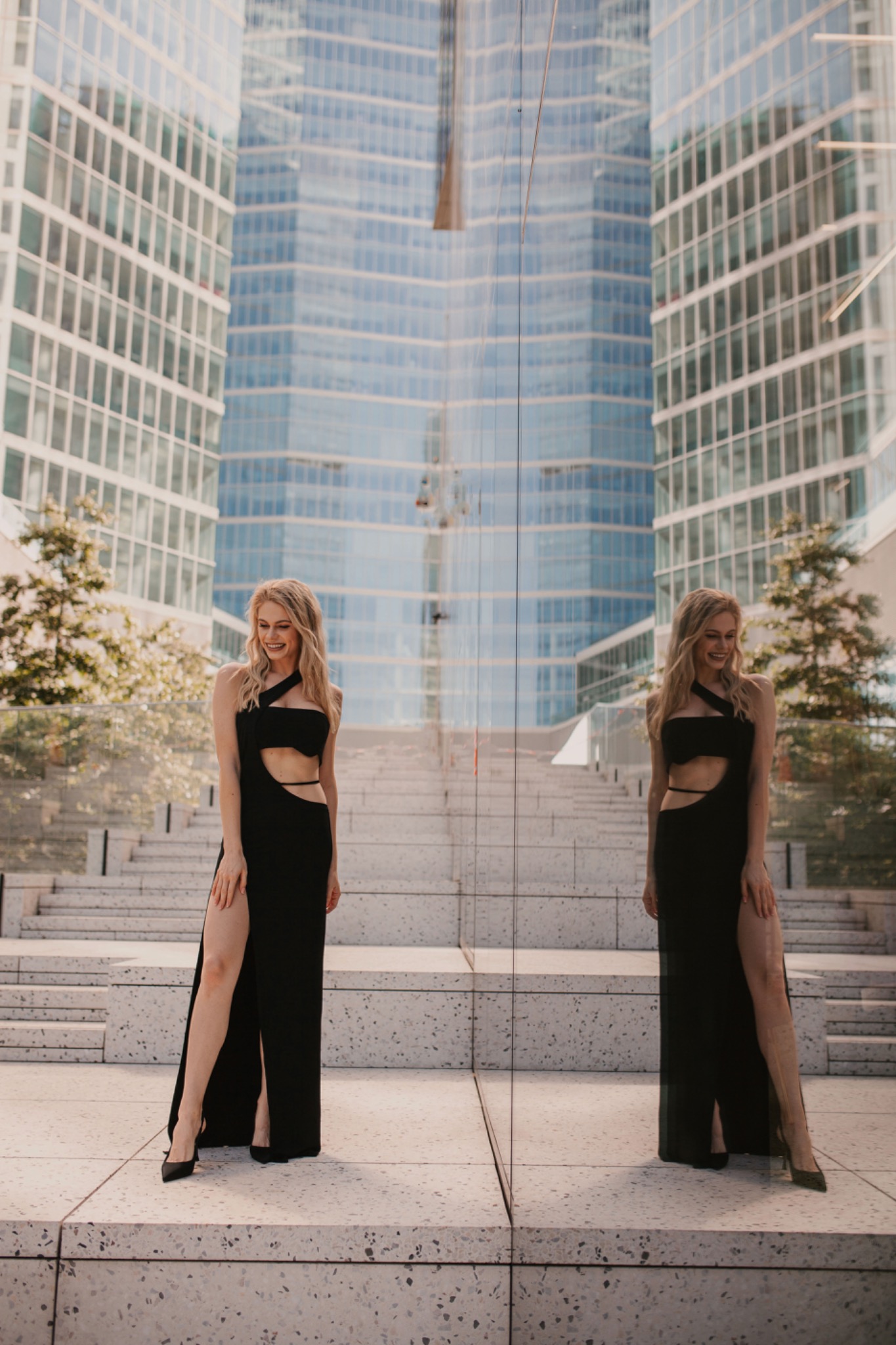 Uśmiechnięta blondynka w czarnej, wyciętej sukni pozuje na tle nowoczesnego budynku, widoczna w odbiciu szklanej ściany.