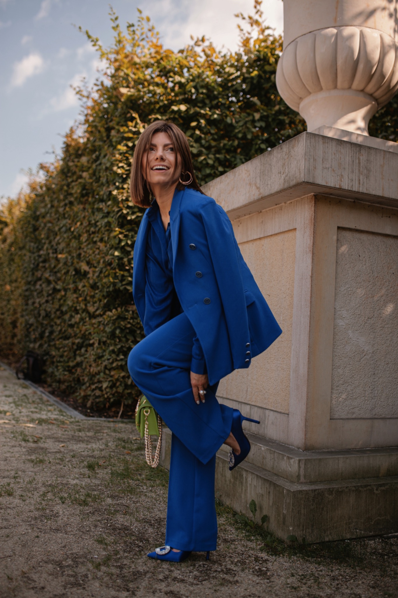 Uśmiechnięta kobieta w kobaltowym garniturze i szpilkach z ozdobną klamrą, pozująca obok kamiennej urny i żywopłotu, z zieloną torebką na złotym łańcuszku.