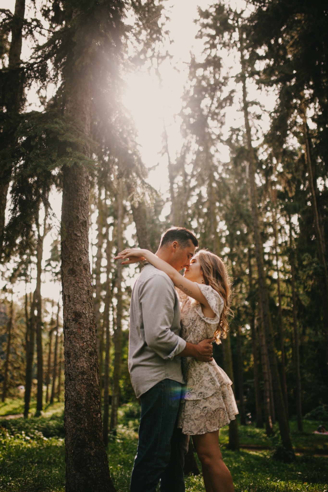 Para całująca się w lesie, promienie słońca przebijają przez korony drzew, ona w krótkiej, wzorzystej sukience, on w szarej koszuli i dżinsach.