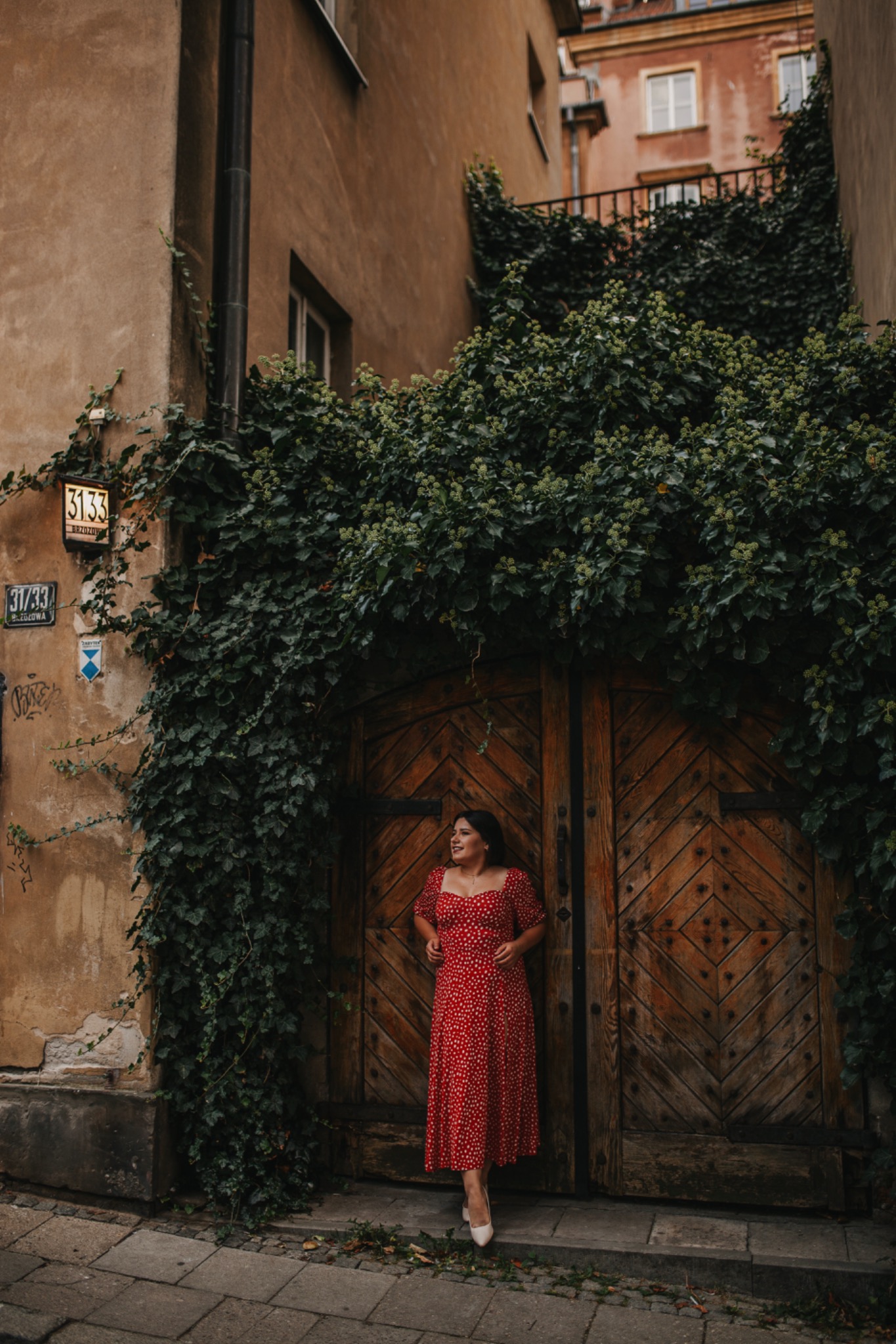 Kobieta w czerwonej sukience w białe groszki pozuje na tle drewnianych drzwi porośniętych bluszczem w Warszawie. Uwagę przykuwa numer 3133 na ścianie budynku po lewej stronie.
