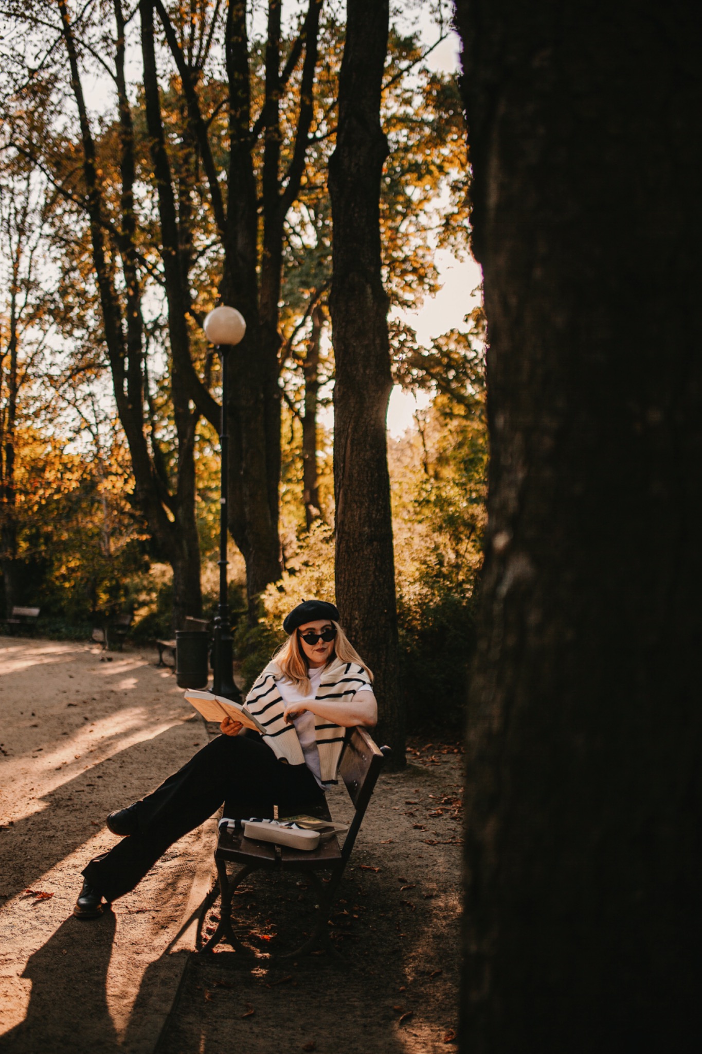 Kobieta w beretce i okularach przeciwsłonecznych siedzi na ławce w parku, czytając książkę w jesiennym słońcu, obok leży pudełko na lunch.