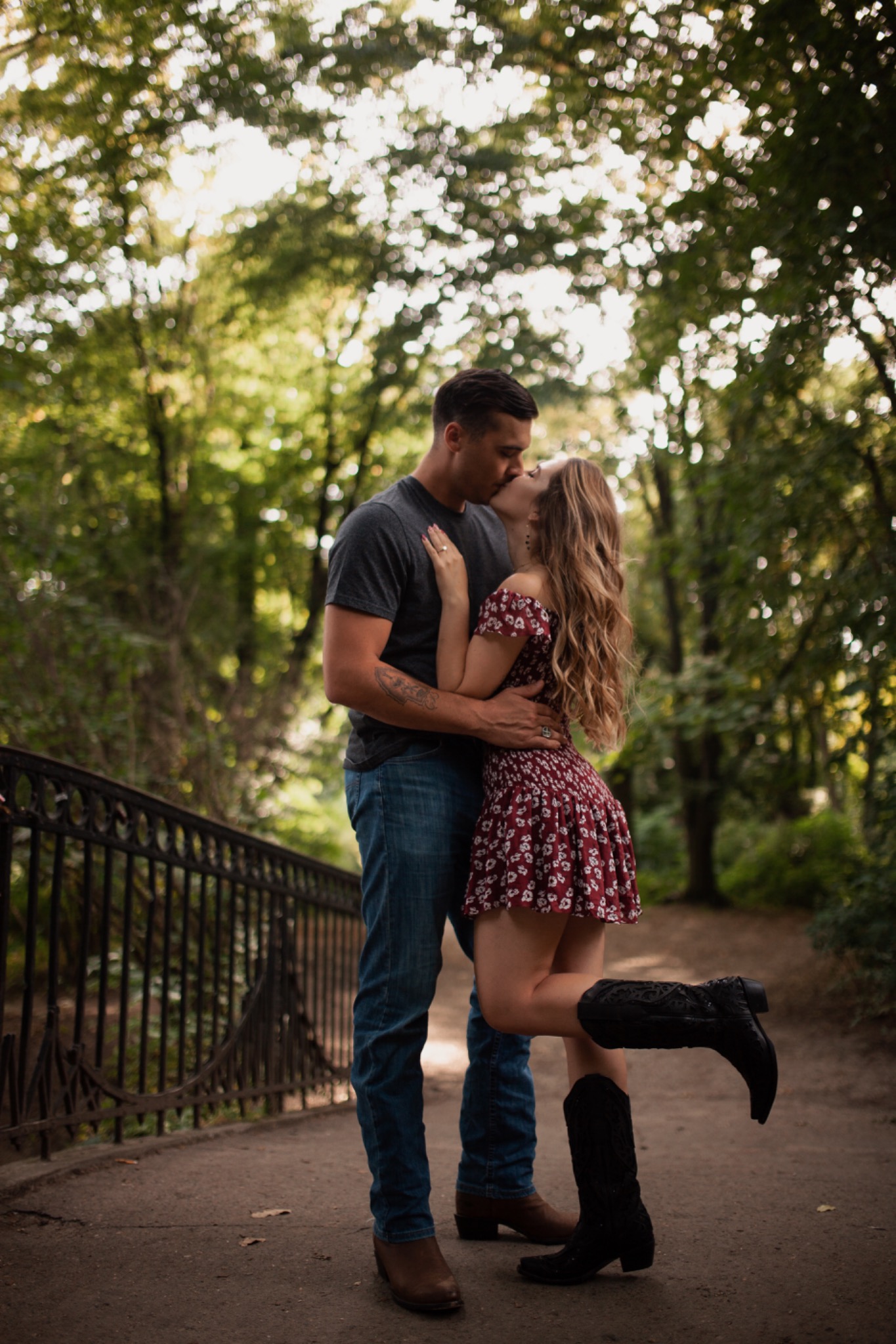 Para całująca się na moście w parku, kobieta w kwiecistej sukience i kowbojkach unosi nogę, mężczyzna w jeansach i szarej koszulce z tatuażem na ramieniu.