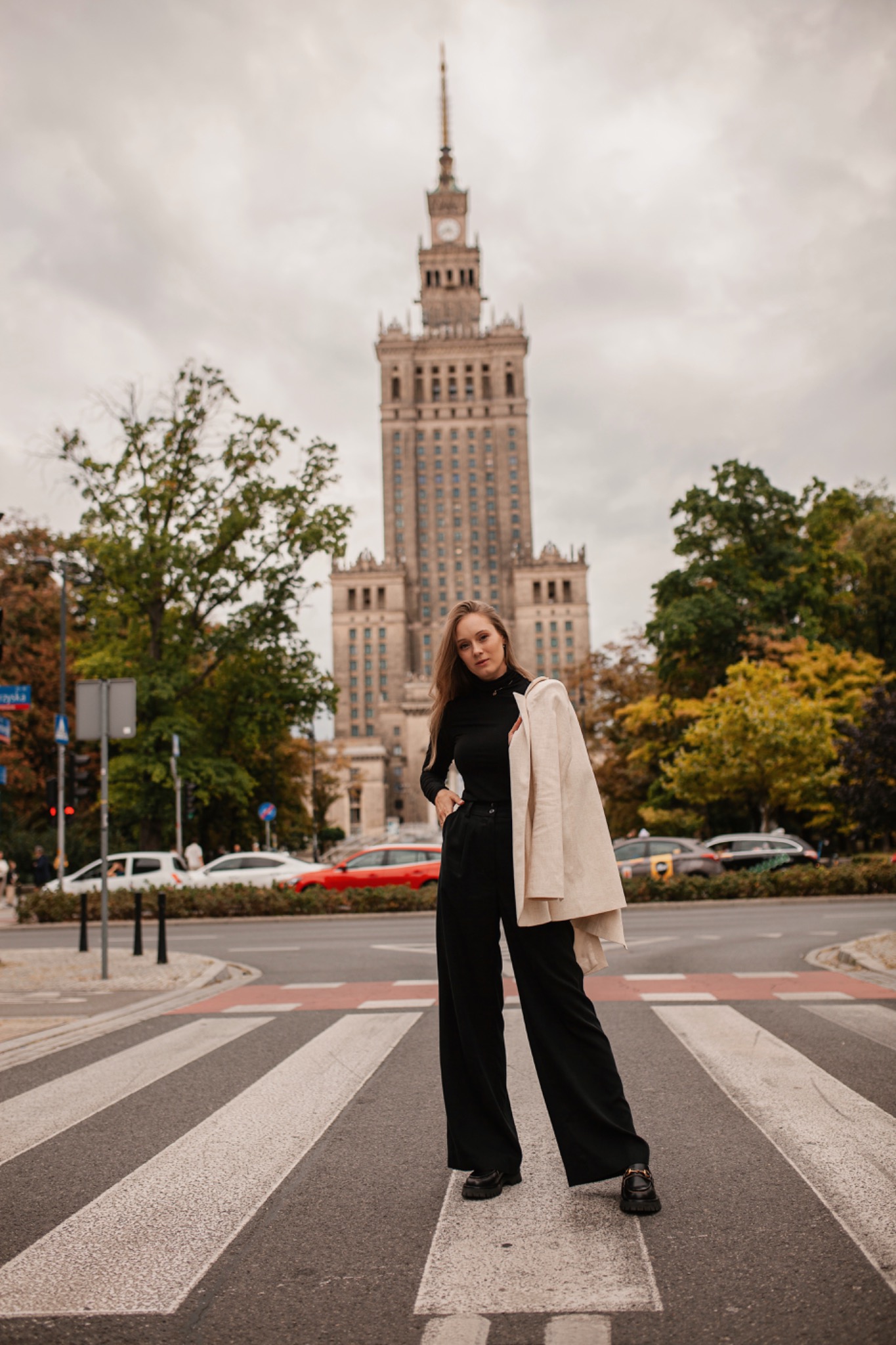 Portret kobiety w czarnym stroju na przejściu dla pieszych, z Pałacem Kultury w tle, jesienna sceneria.