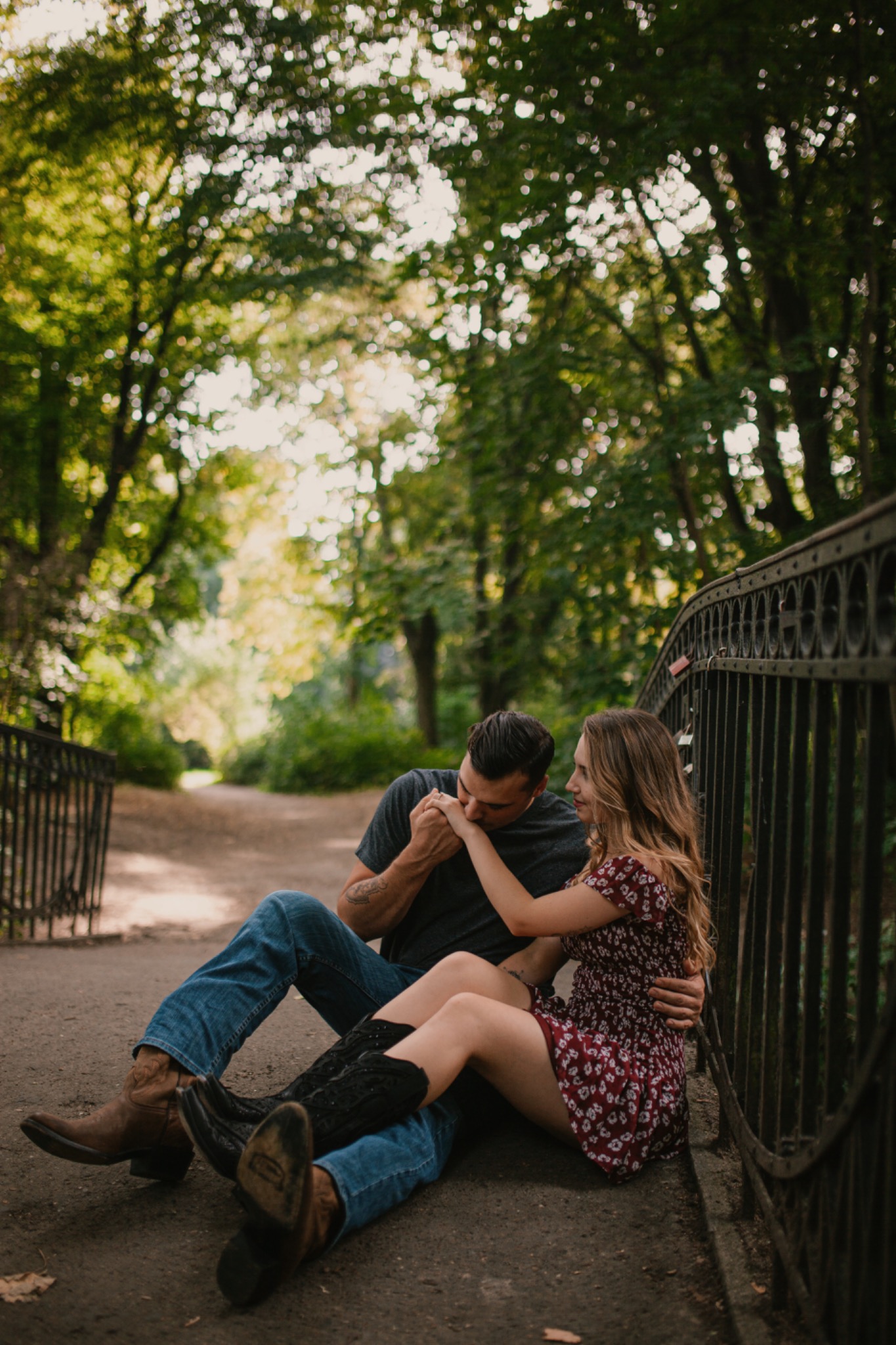 Para siedząca na moście w parku, mężczyzna w kowbojkach całuje dłoń kobiety w kwiecistej sukience, w tle zieleń drzew i ścieżka.