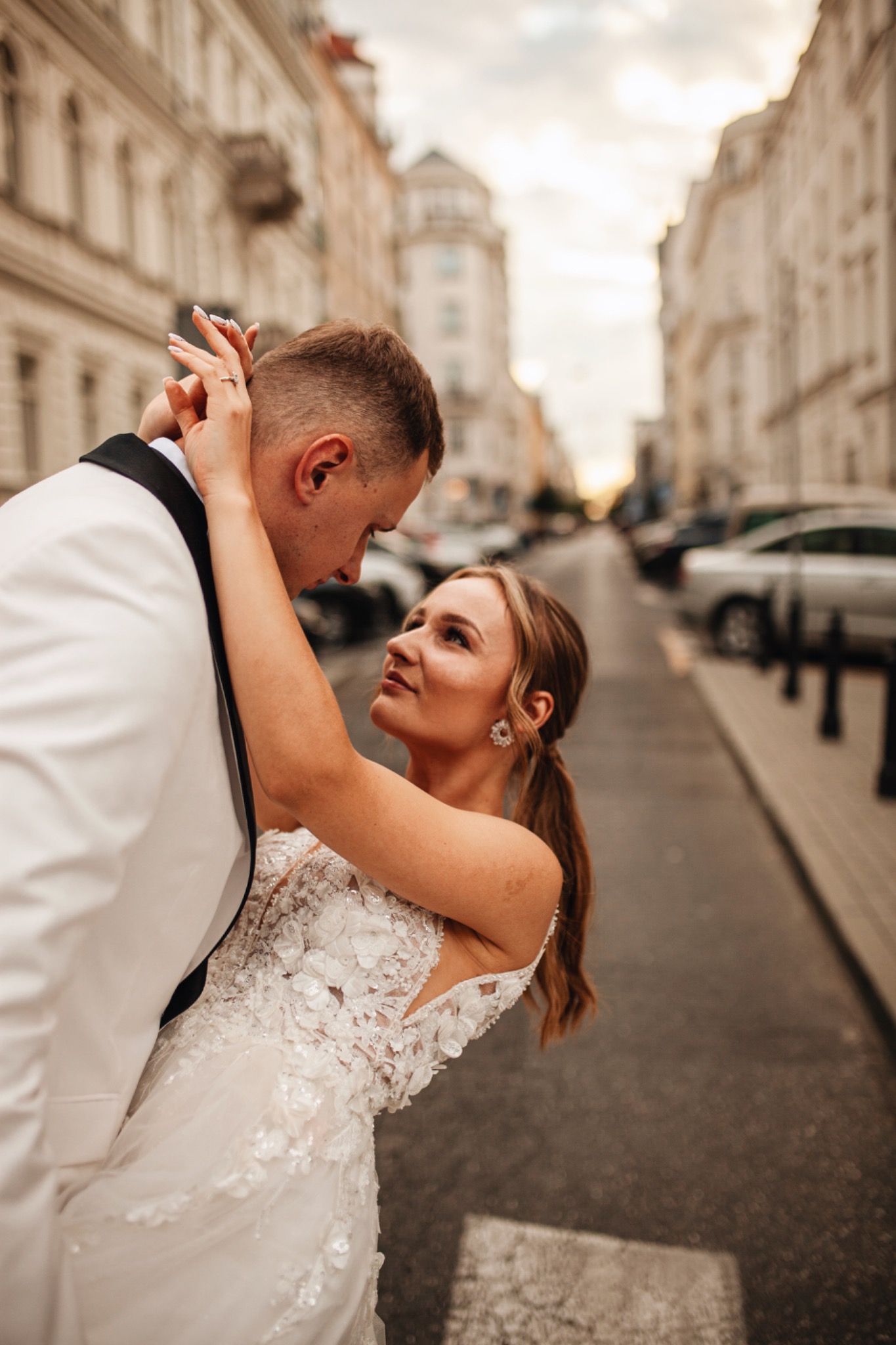 Panna młoda w koronkowej sukni i pan młody w białym smokingu obejmujący się na tle miejskiej ulicy z perspektywy żabiej. Pierścionek zaręczynowy na palcu panny młodej.