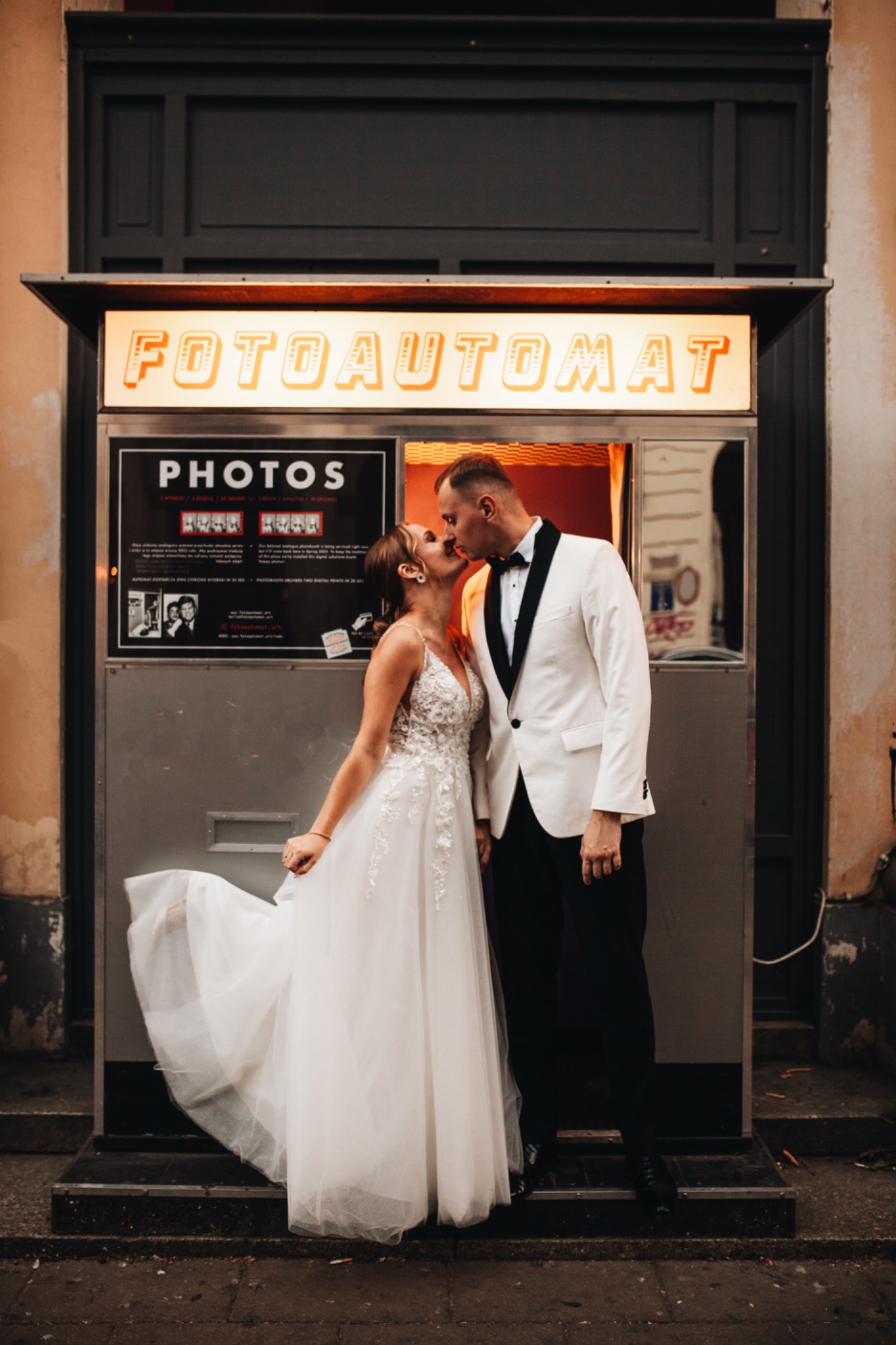 Pocałunek nowożeńców przed budką fotoautomat. Panna młoda w białej sukni z koronkową górą, pan młody w białym smokingu z czarną muchą. Nad nimi napis FOTOAUTOMAT.