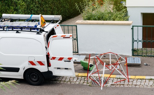 Biały furgon z otwartymi drzwiami, znak ostrzegawczy, barierka i studzienka kanalizacyjna sugerują prace instalatorskie w otoczeniu zieleni i ogrodzenia posesji.