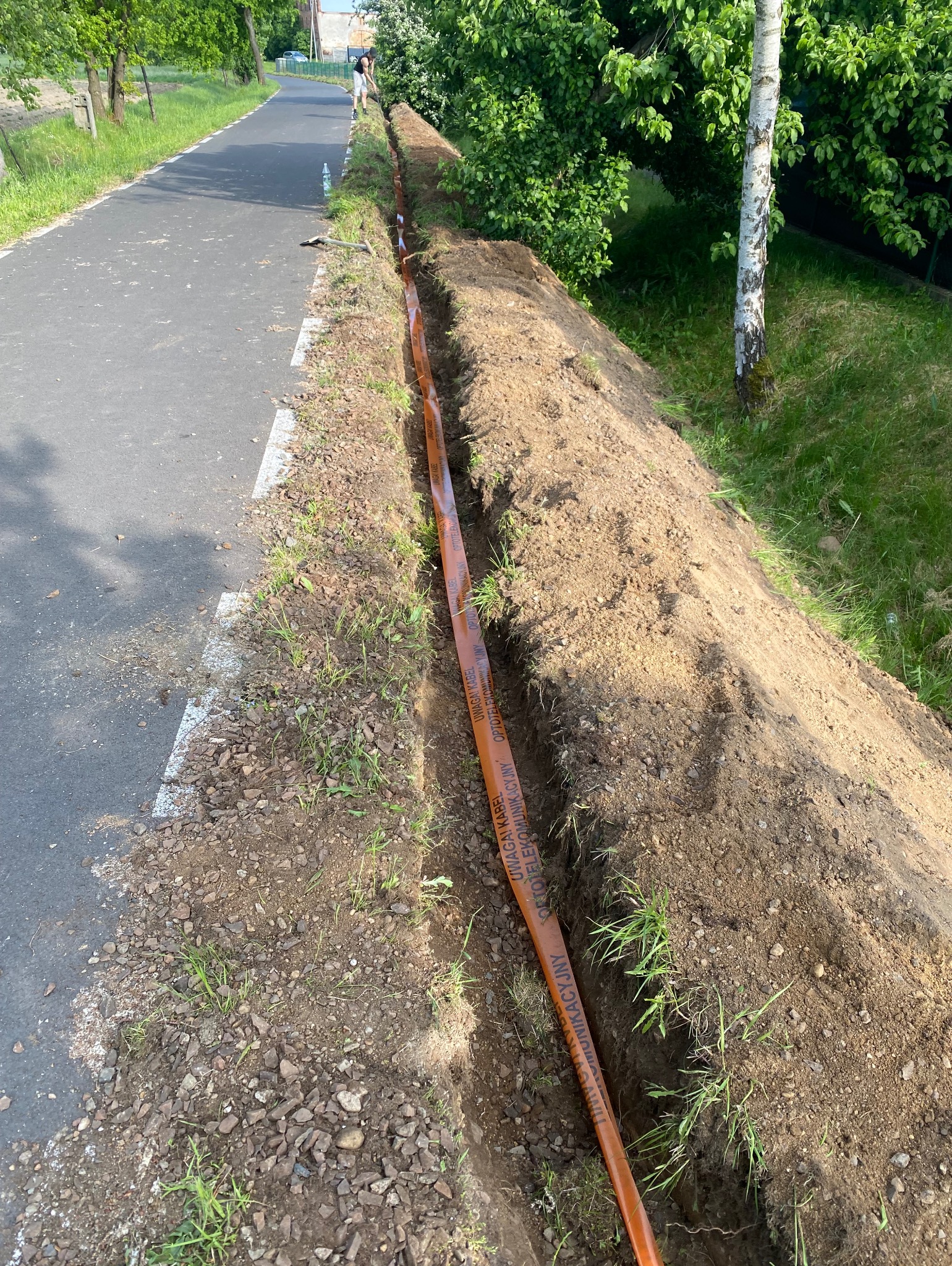 Wkopany pomarańczowy kabel telekomunikacyjny wzdłuż drogi asfaltowej, widok z góry na wykop, częściowo zasypany ziemią, w tle drzewa i osoba.