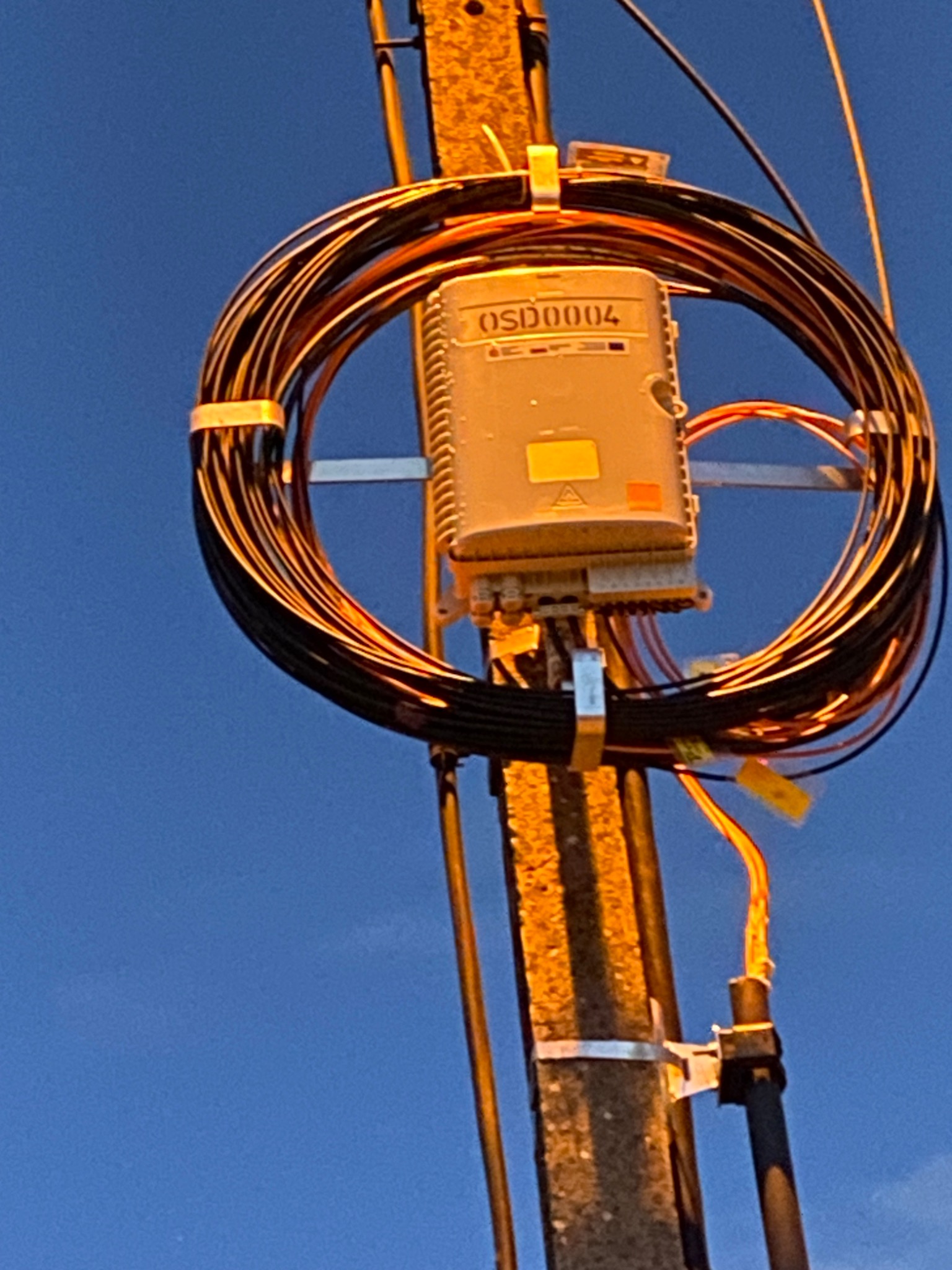 Słup telekomunikacyjny z zamontowaną skrzynką OSD0004 i kablem światłowodowym zwiniętym w okrąg, zabezpieczonym opaskami. Błękitne tło nieba.