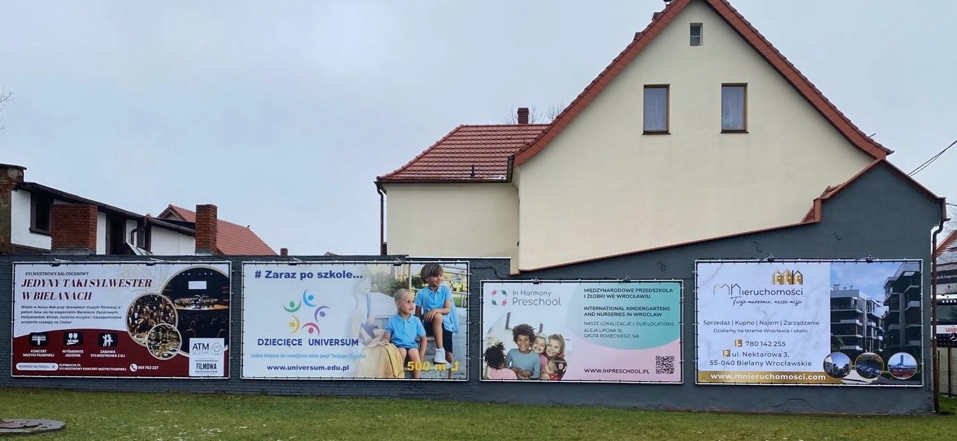 Rząd czterech banerów reklamowych na szarym murze, z budynkami w tle. Banery promują wydarzenie sylwestrowe, przedszkole, żłobek i agencję nieruchomości.