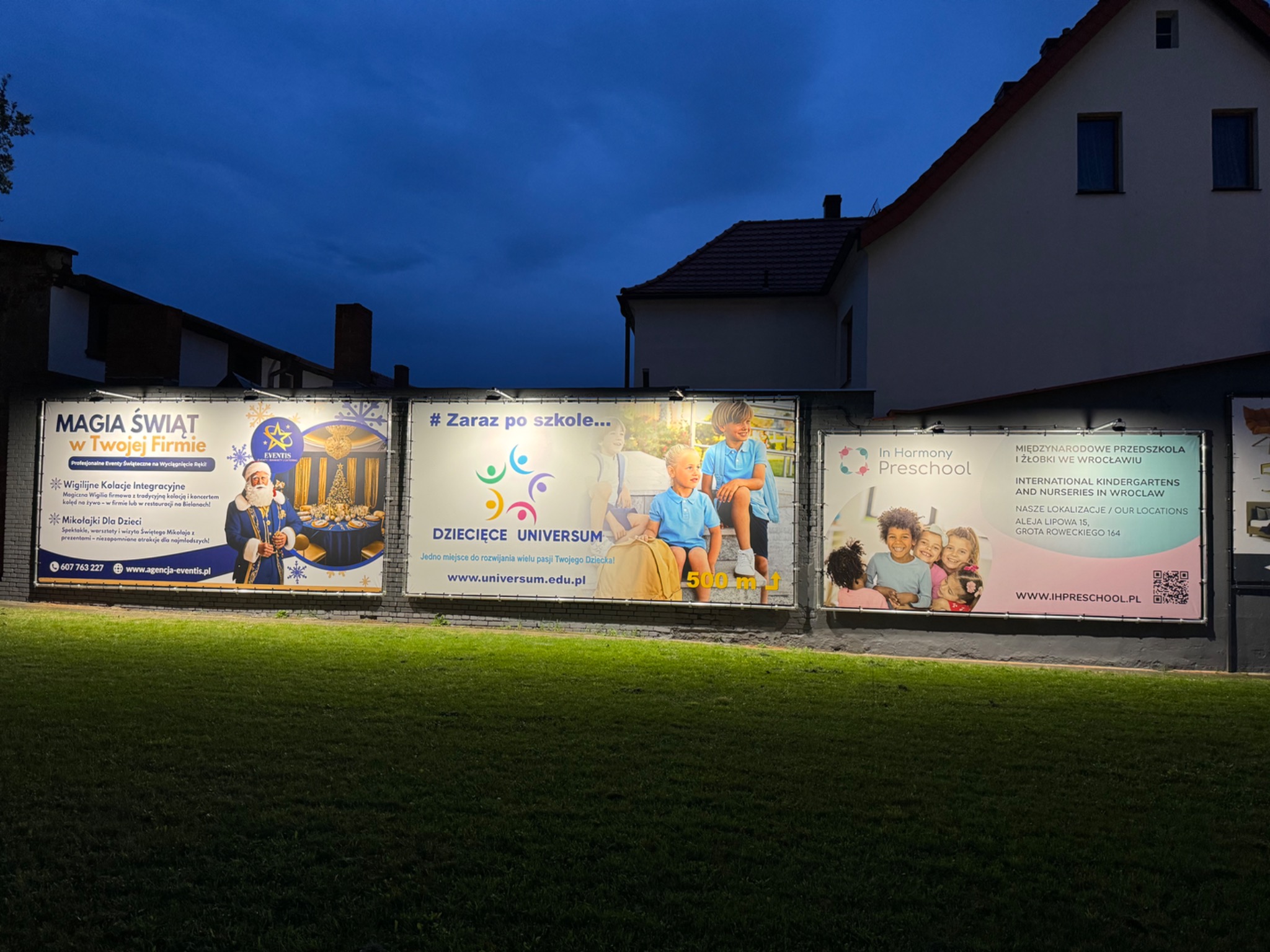 Instalacja trzech podświetlonych billboardów reklamowych z różnymi treściami (eventy, przedszkole) na tle budynku i trawnika, oświetlonych nocą.