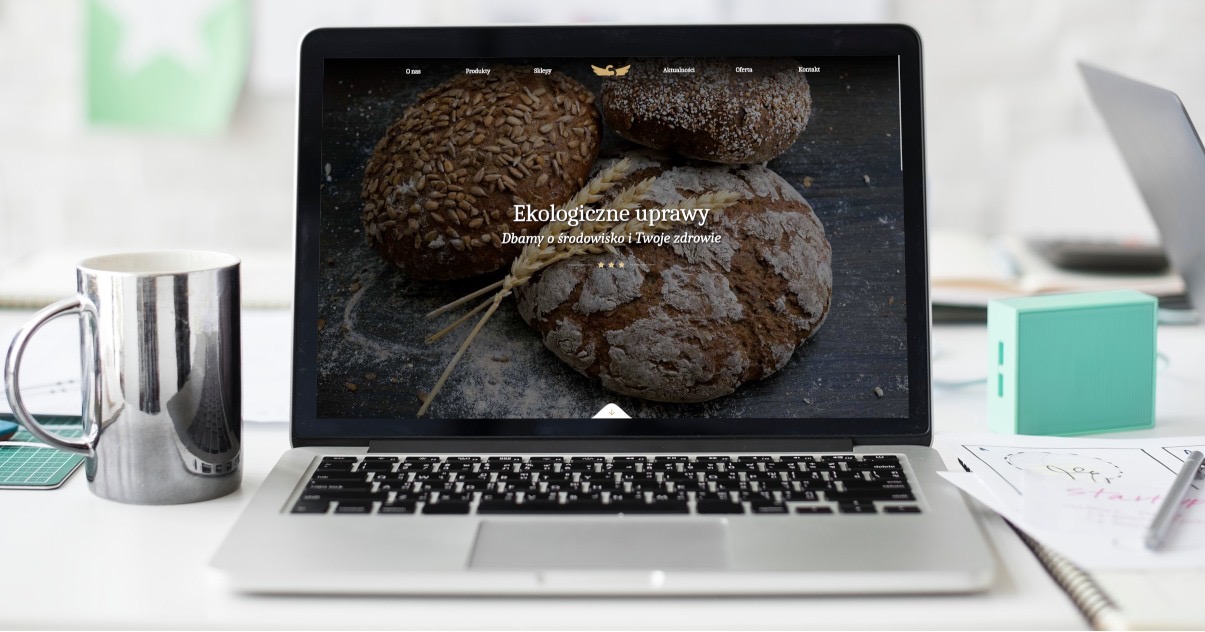Laptop wyświetla stronę internetową z motywem piekarniczym: trzy bochenki chleba, napis 'Ekologiczne uprawy'. Na biurku srebrny kubek, turkusowy powerbank i szkice interfejsu.