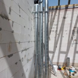 INVEST DL SPÓŁKA Z OGRANICZONĄ ODPOWIEDZIALNOŚCIĄ - Montaż pionowych, spiralnych rur wentylacyjnych w surowym wnętrzu budynku z białych bloczków, widoczne narzędzia i akcesoria montażowe.