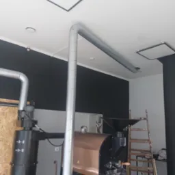 System wentylacyjny z metalowymi rurami odprowadzającymi powietrze od pieca do palenia kawy, widoczna drabina oparta o ścianę.