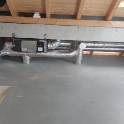 INVEST DL SPÓŁKA Z OGRANICZONĄ ODPOWIEDZIALNOŚCIĄ - Instalacja rekuperacji w surowym stanie poddasza: srebrne rury izolowane termicznie, centrala wentylacyjna zamontowana pod drewnianą konstrukcją dachu.