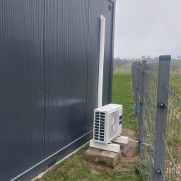 INVEST DL SPÓŁKA Z OGRANICZONĄ ODPOWIEDZIALNOŚCIĄ - Zewnętrzna jednostka klimatyzatora zamontowana przy metalowym kontenerze, widoczny biały przewód odprowadzający skropliny, szara siatka ogrodzeniowa i trawnik z rozrzuconymi śmieciami w tle.