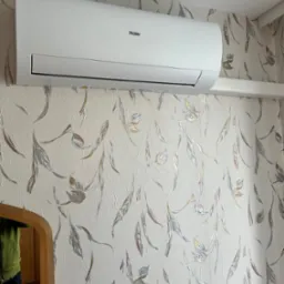 Biały klimatyzator Haier zamontowany na ścianie z tapetą w liście. Widoczne odbicie w lustrze. Ujęcie z dołu, podkreśla estetykę urządzenia i dopasowanie do wnętrza.