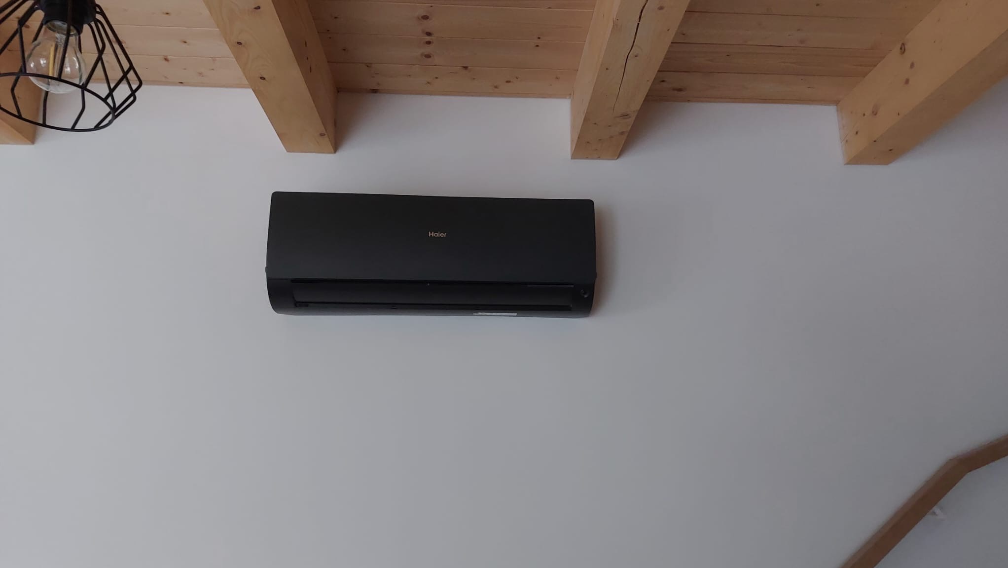 Czarny klimatyzator Haier zamontowany na białej ścianie w pomieszczeniu z drewnianym stropem i nowoczesną lampą wiszącą.