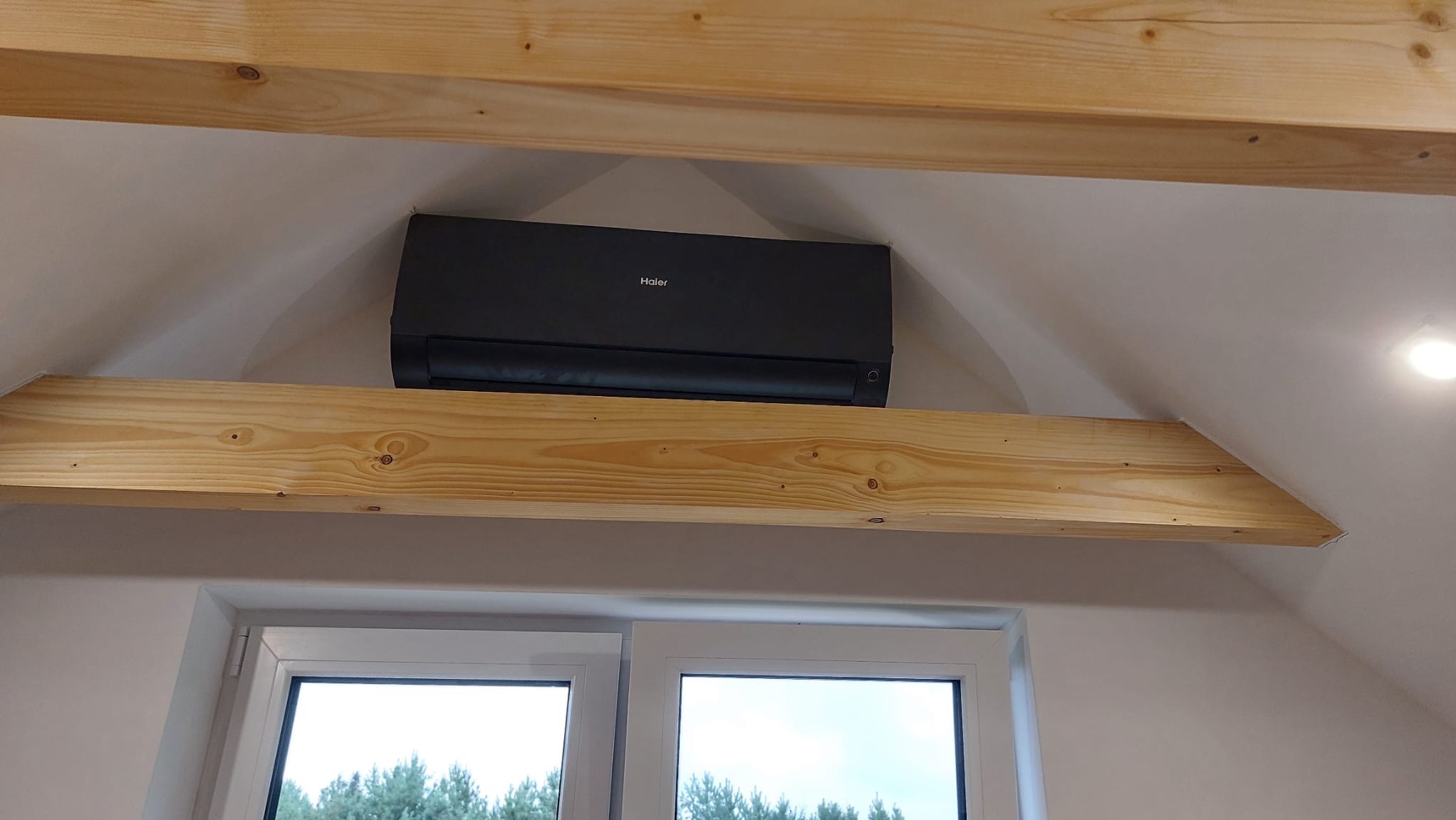 Czarna jednostka klimatyzacji Haier zamontowana pod drewnianą belką na poddaszu, widok z dołu z oknem w tle.