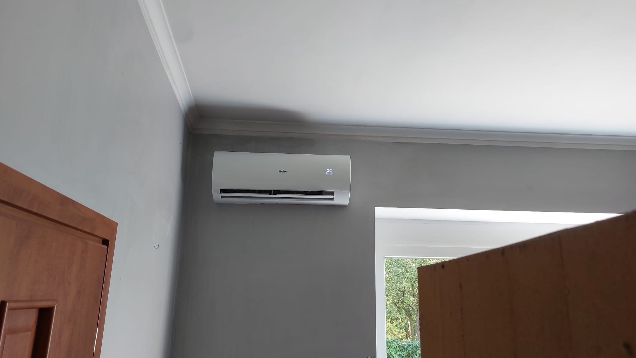 Biała jednostka klimatyzacyjna Haier, z wyświetlaczem temperatury 25 stopni Celsjusza, zamontowana na szarej ścianie w pomieszczeniu w Giżycku, widok z boku, z fragmentem drewnianych drzwi po lewej...