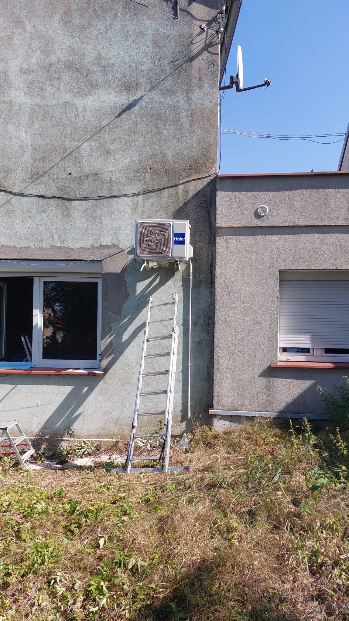 Zewnętrzna jednostka klimatyzatora Haier zamontowana na szarej ścianie budynku, z opartą o nią aluminiową drabiną, widoczne okno i zaniedbane otoczenie z chwastami.