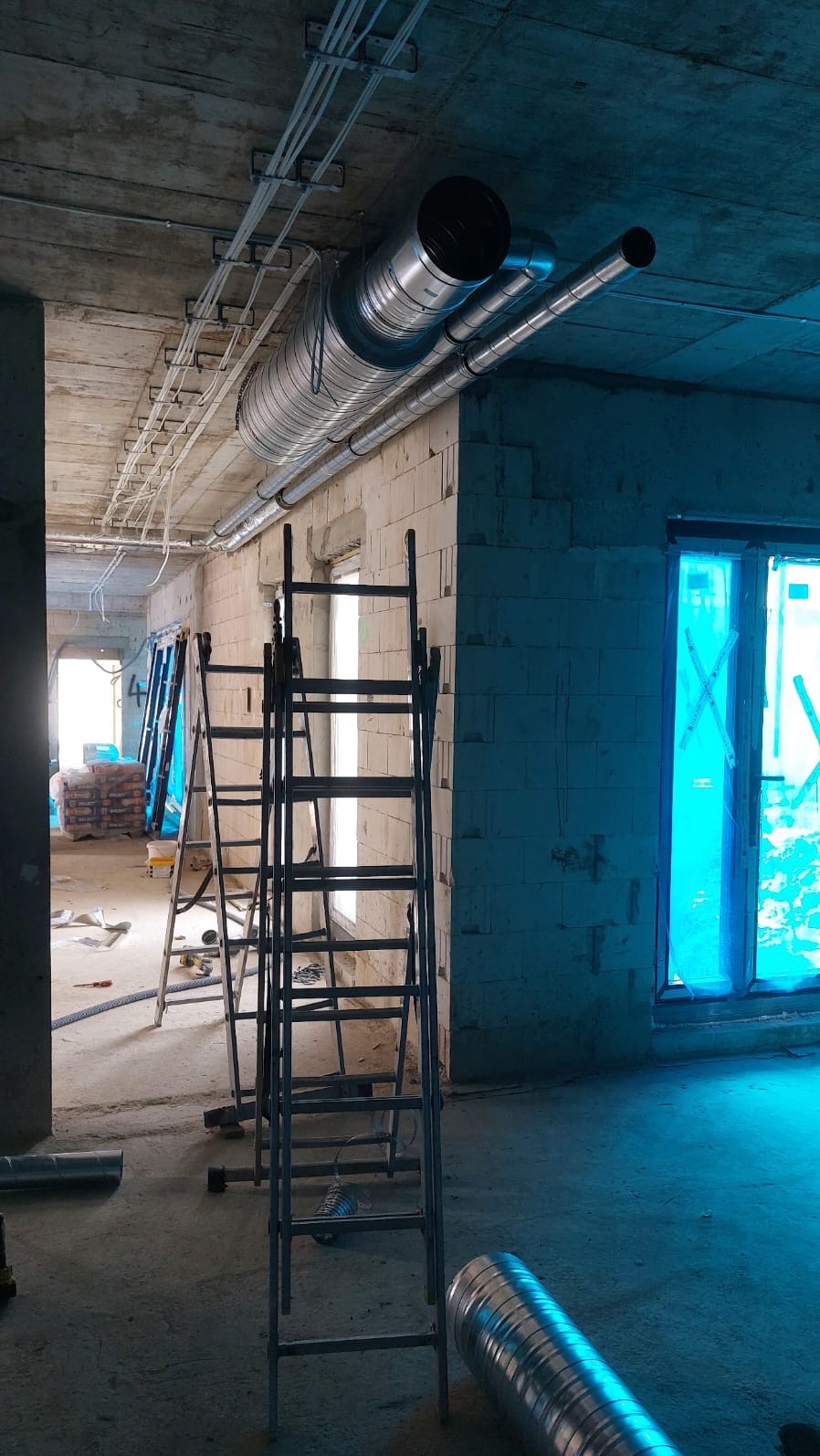 Wnętrze w stanie surowym z widocznymi rurami wentylacyjnymi pod sufitem, drabiny opierają się o ścianę z oknami zabezpieczonymi folią.