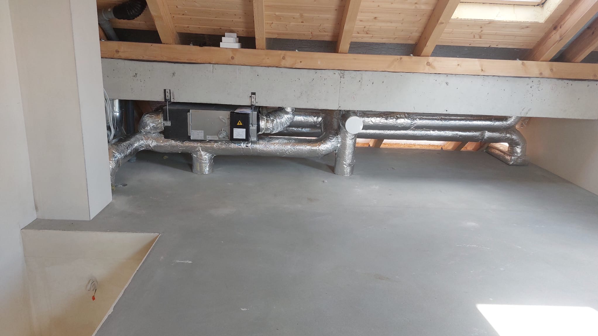 Instalacja rekuperacji w surowym stanie poddasza: srebrne rury izolowane termicznie, centrala wentylacyjna zamontowana pod drewnianą konstrukcją dachu.