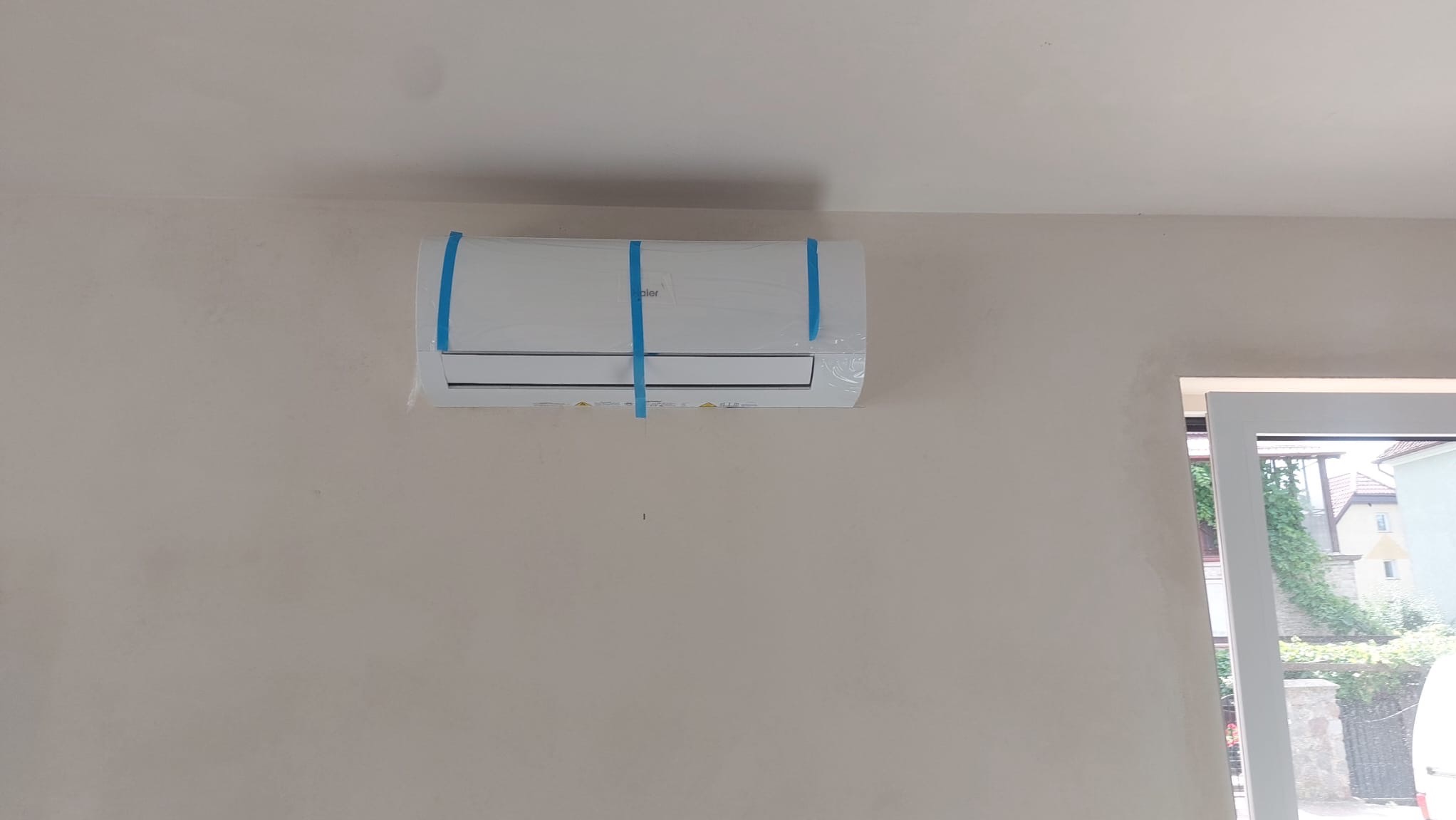 Nowy, biały klimatyzator marki Haier, zabezpieczony niebieską taśmą, zamontowany na beżowej ścianie w pokoju, widoczne okno z widokiem na zewnątrz.
