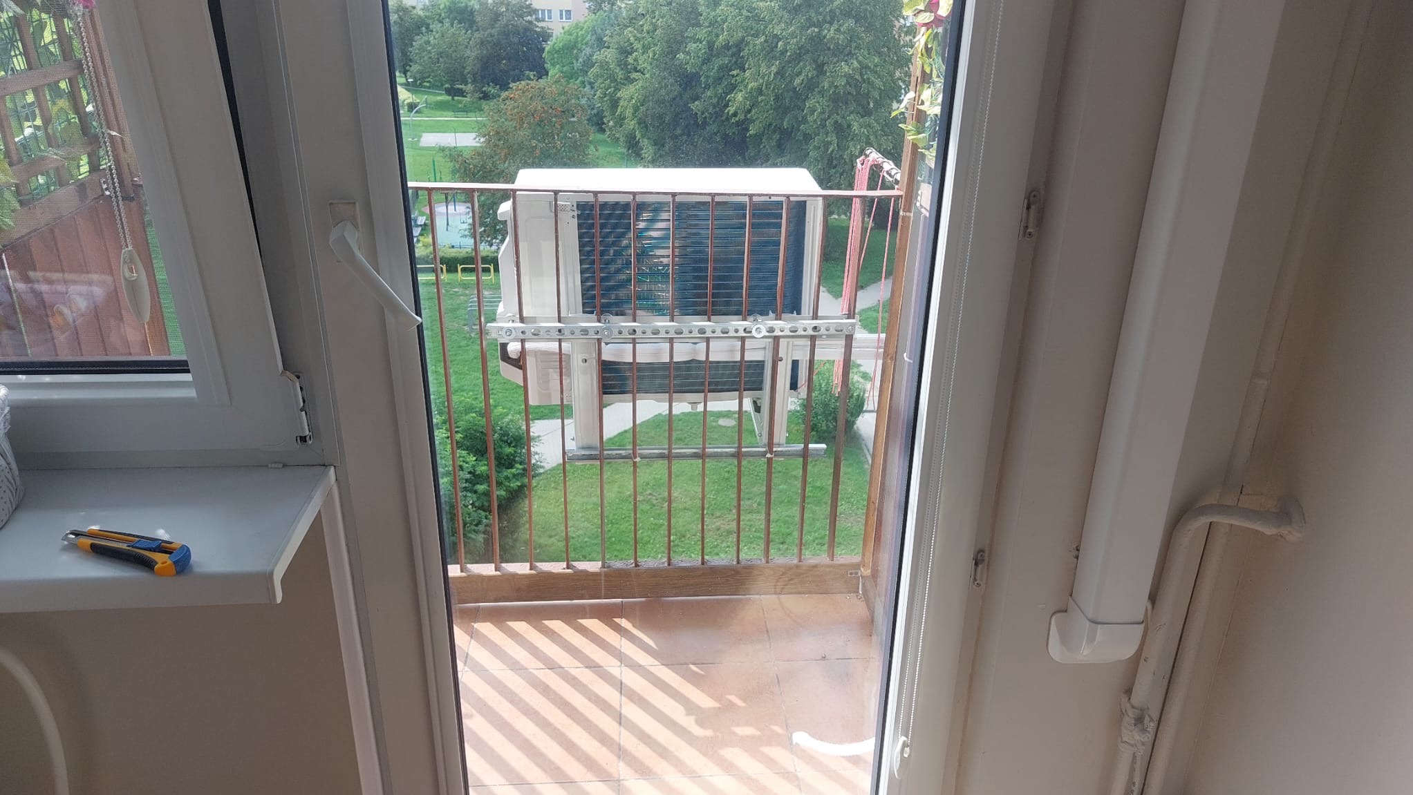 Widok z balkonu na zewnętrzną jednostkę klimatyzatora zamontowaną na balustradzie, z narzędziem - nożykiem na parapecie.