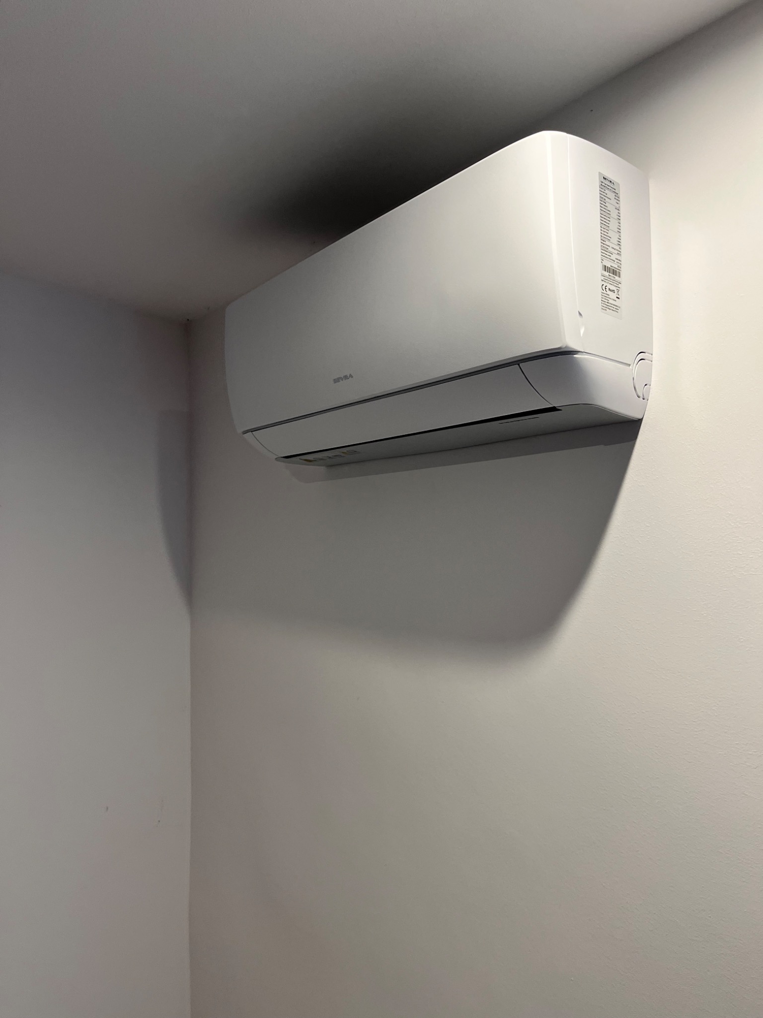 Biała jednostka wewnętrzna klimatyzacji zamontowana na jasnoszarej ścianie w rogu pomieszczenia, widoczne detale obudowy i naklejki informacyjne.