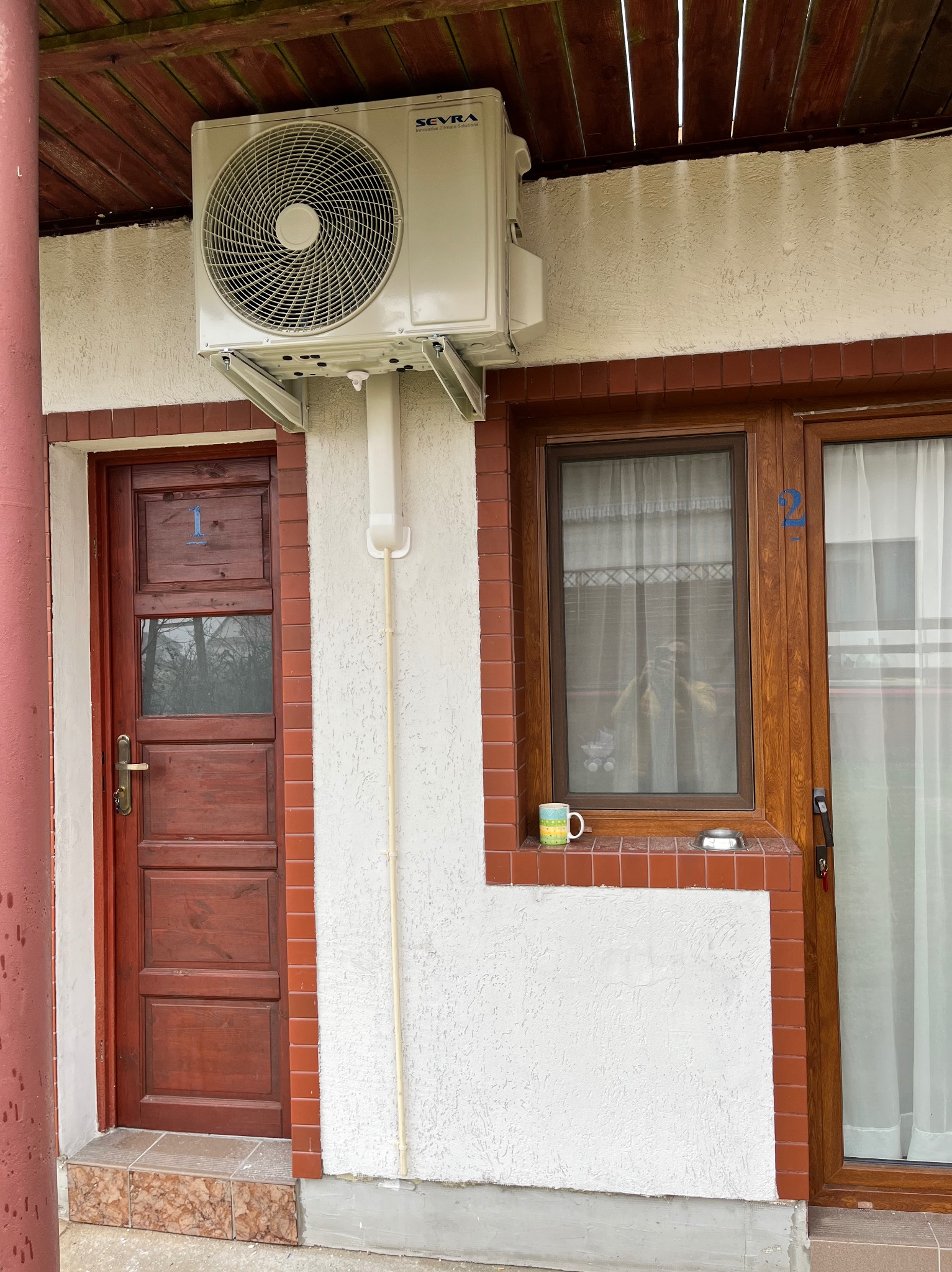 Zewnętrzna jednostka klimatyzatora marki Sevra zamontowana nad drzwiami oznaczonymi numerem 1 i oknem oznaczonym numerem 2, z kubkiem i popielniczką na parapecie.