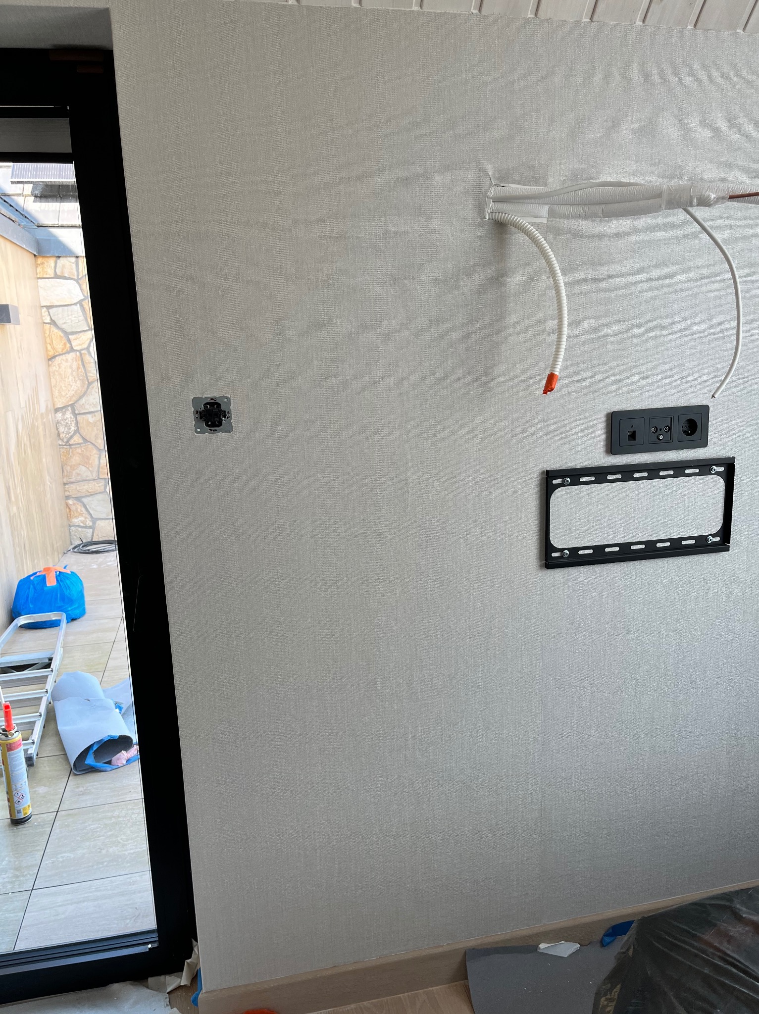 Przygotowanie do montażu klimatyzacji w domu: widoczne otwory na kable, gniazdka elektryczne, uchwyt naścienny, przewody w peszlu, ściana pokryta tapetą o delikatnej strukturze, w tle przez okno...