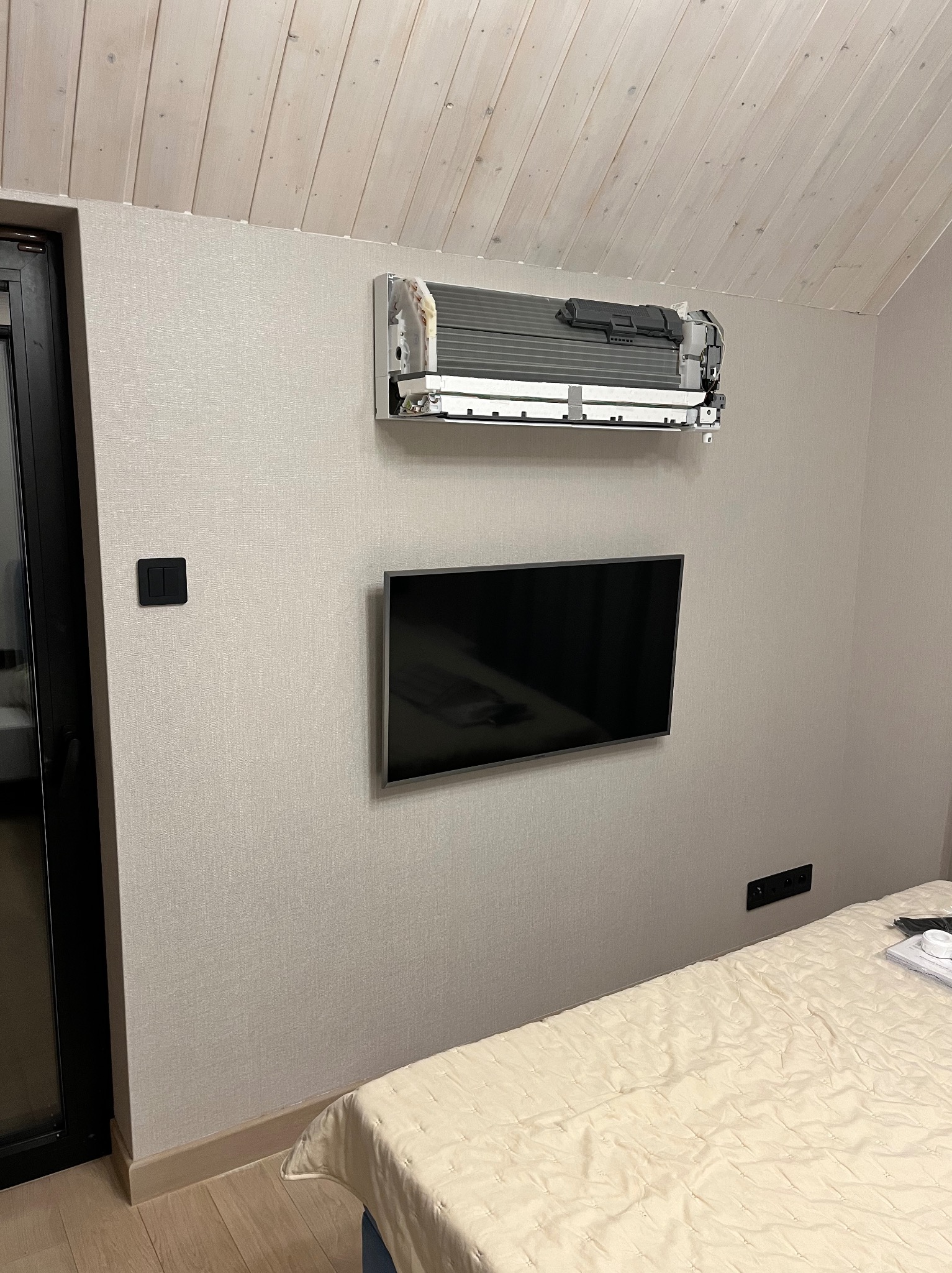 Wnętrze sypialni z zamontowaną, ale częściowo rozebraną jednostką klimatyzatora na ścianie nad telewizorem i łóżkiem.