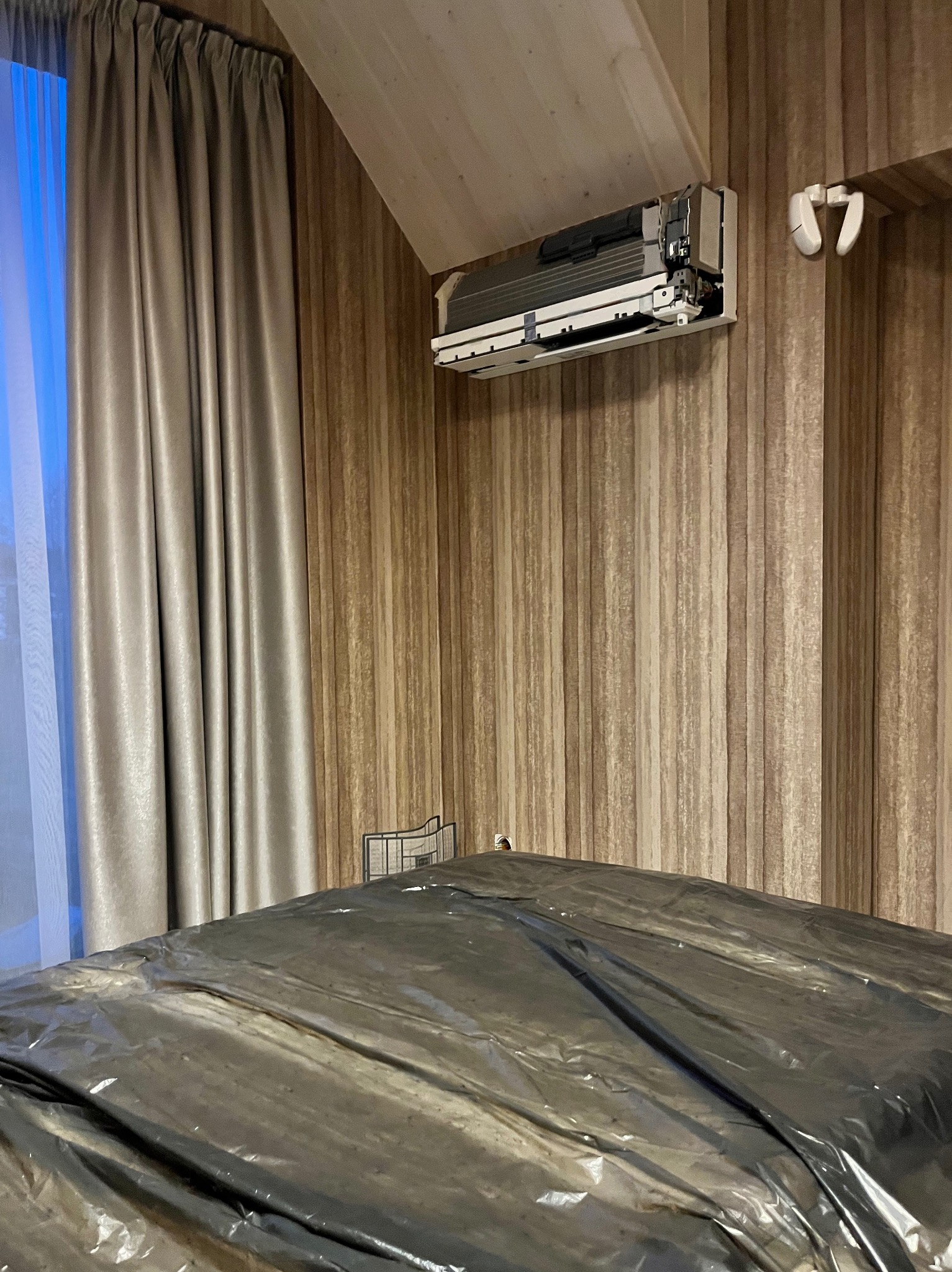 Rozłożona folia ochronna na łóżku w pomieszczeniu z zamontowaną klimatyzacją ścienną, widoczne otwarte wnętrze urządzenia, w tle zasłony i tapeta w pionowe pasy.