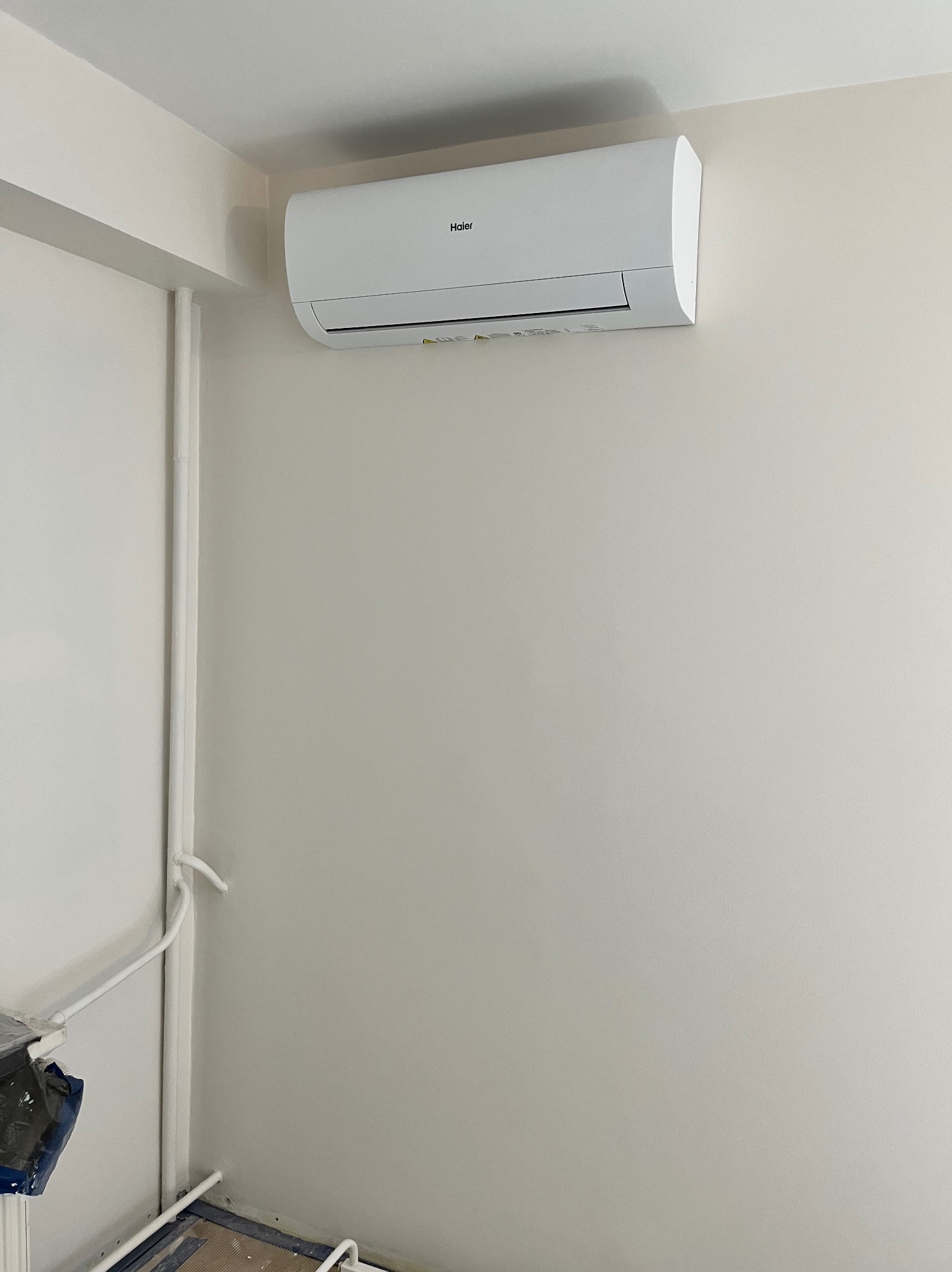 Biały klimatyzator marki Haier zamontowany na jasnej ścianie w rogu pomieszczenia, widoczne rury przy ścianie po lewej stronie.