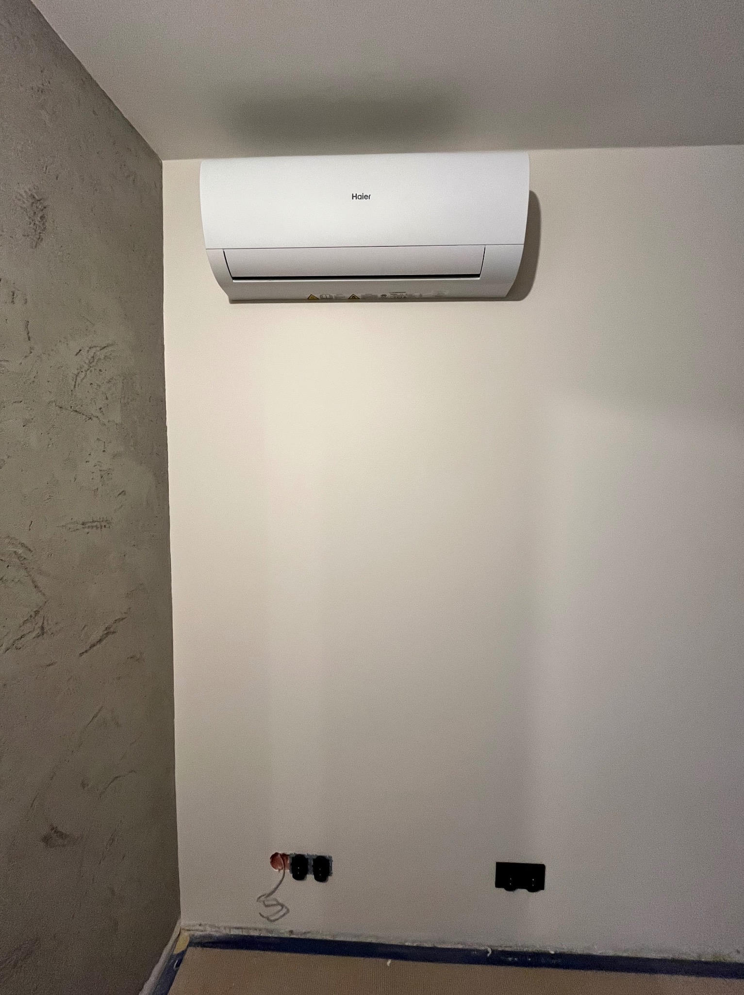 Biała klimatyzacja Haier zamontowana na ścianie w pokoju, widoczne gniazdka elektryczne pod klimatyzatorem.