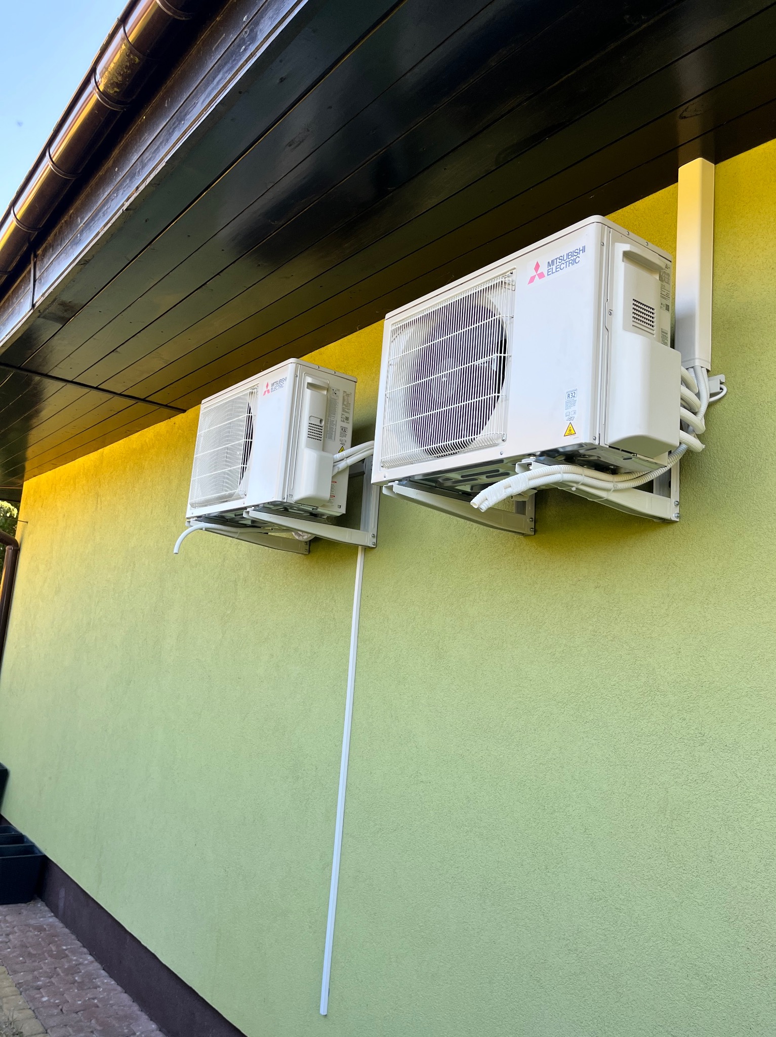 Dwa klimatyzatory Mitsubishi Electric zamontowane na zielonej ścianie domu w Giżycku, widoczne rury i przewody instalacyjne.